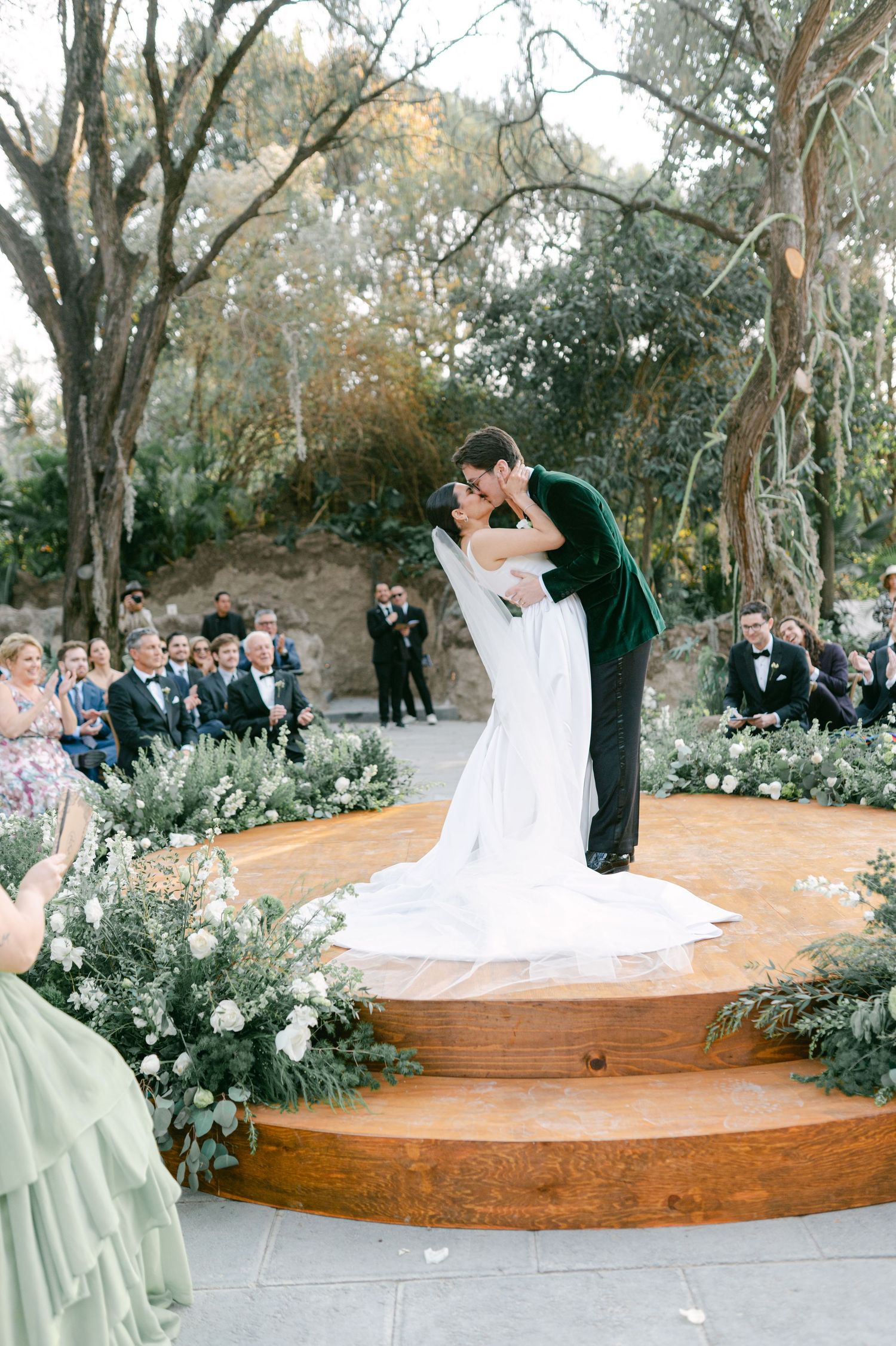 Bride and groom sharing first kiss on circular wooden altar surrounded by lush greenery at outdoor wedding.