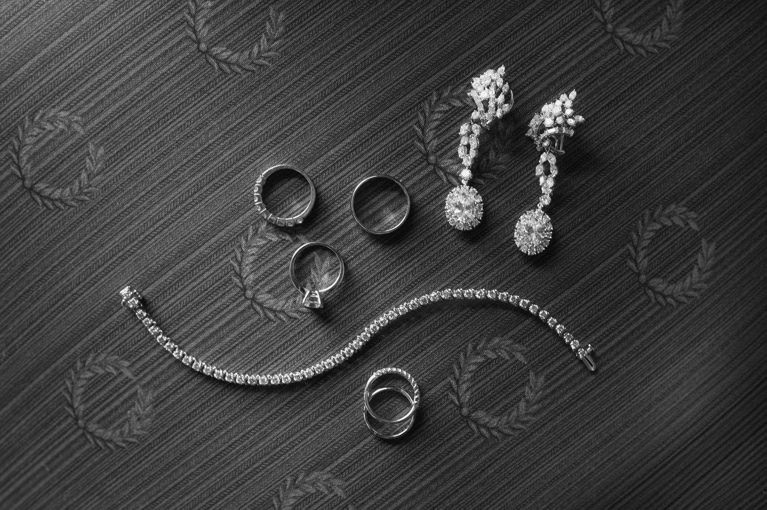 Black and white flat lay of wedding jewelry including diamond earrings, rings, and tennis bracelet on dark surface.