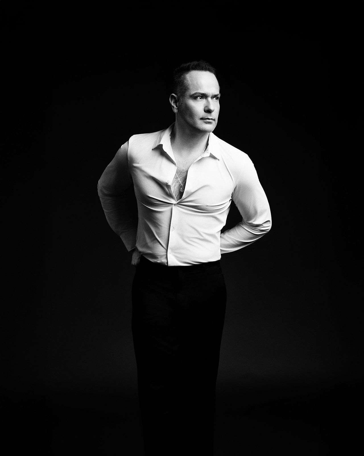 Black and white studio portrait of a man in white shirt and black trousers posing confidently against dark background.