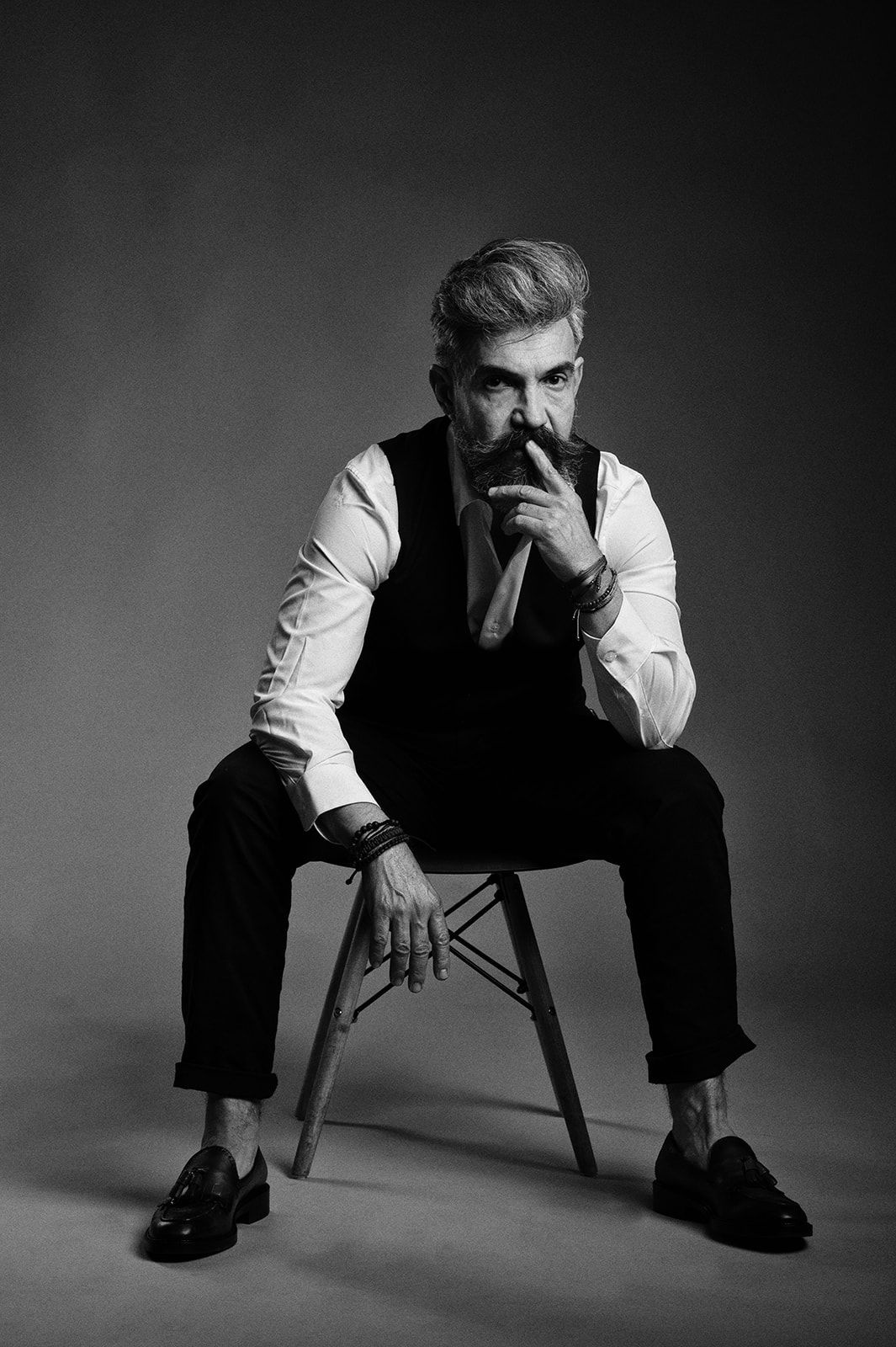Black and white full body portrait of a bearded man in vest and white shirt seated on modern chair.