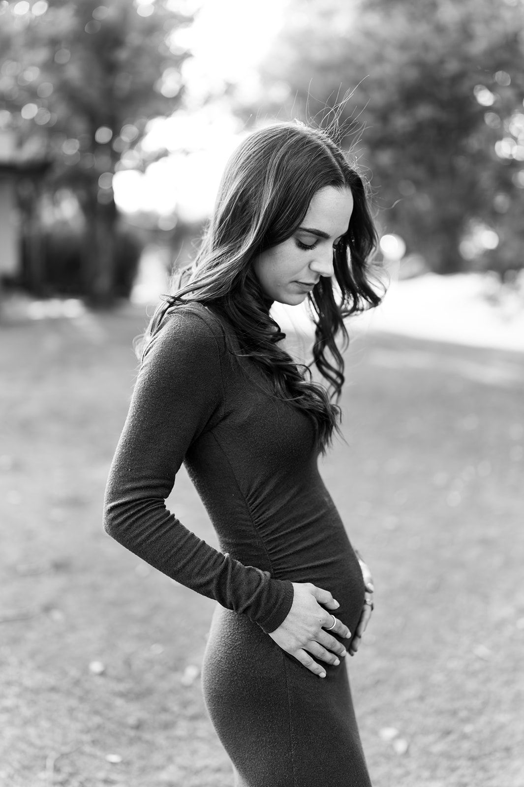 Black and white portrait of pregnant woman in gray dress cradling baby bump outdoors.