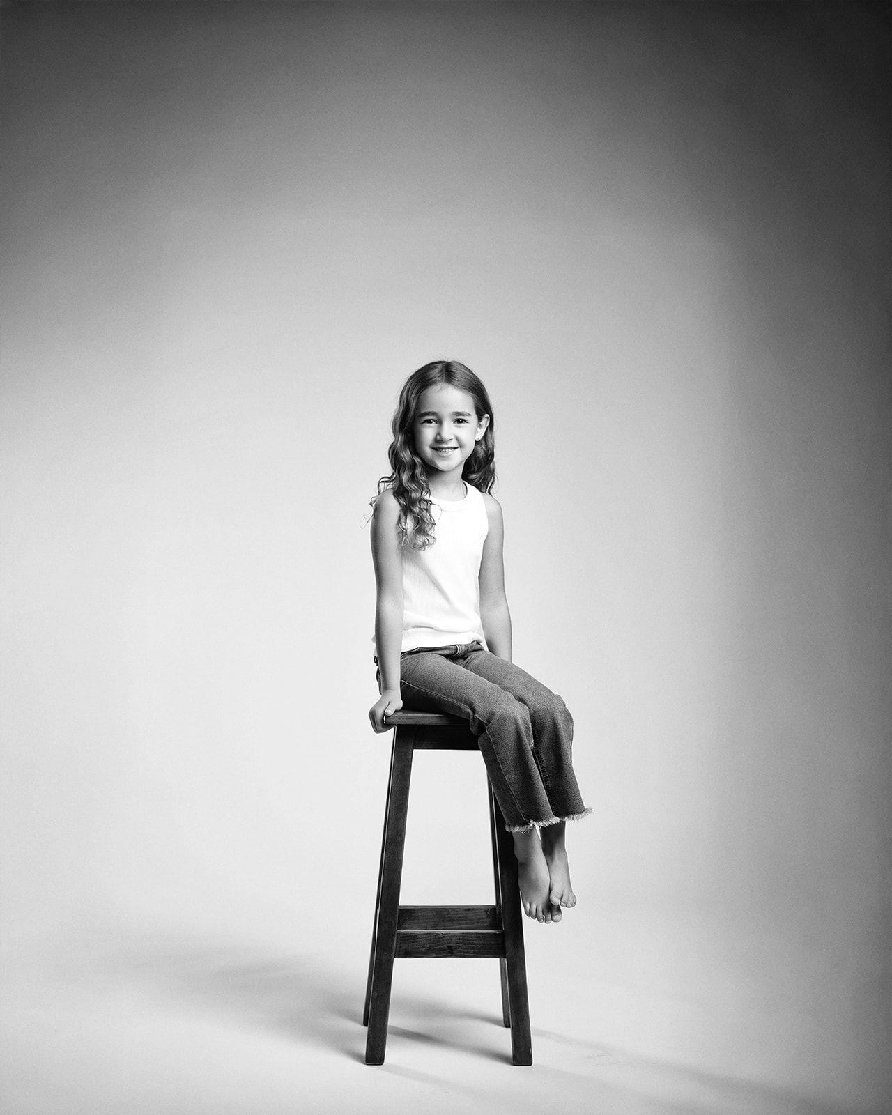 Full-length black and white studio portrait of a young girl sitting on a tall wooden stool, smiling.