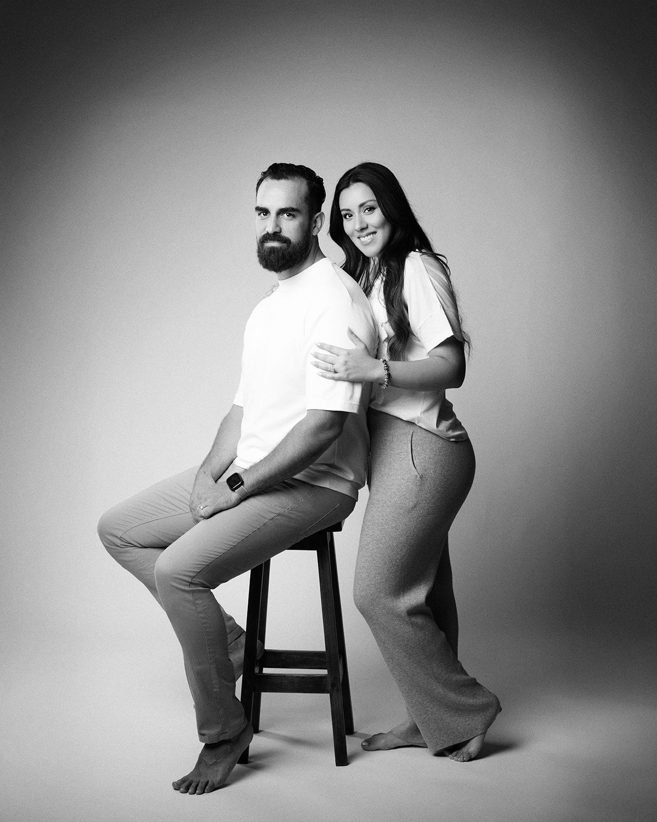 Black and white studio portrait of a bearded man sitting on a stool and a woman leaning against his back.