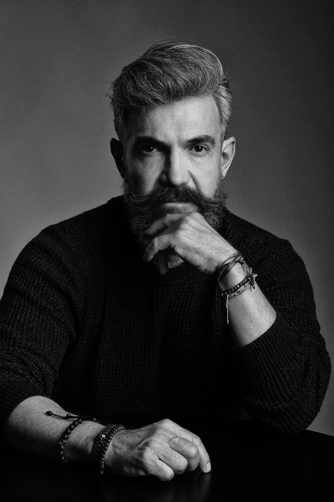 Black and white close-up portrait of a bearded man in dark sweater with bracelets, hands clasped.