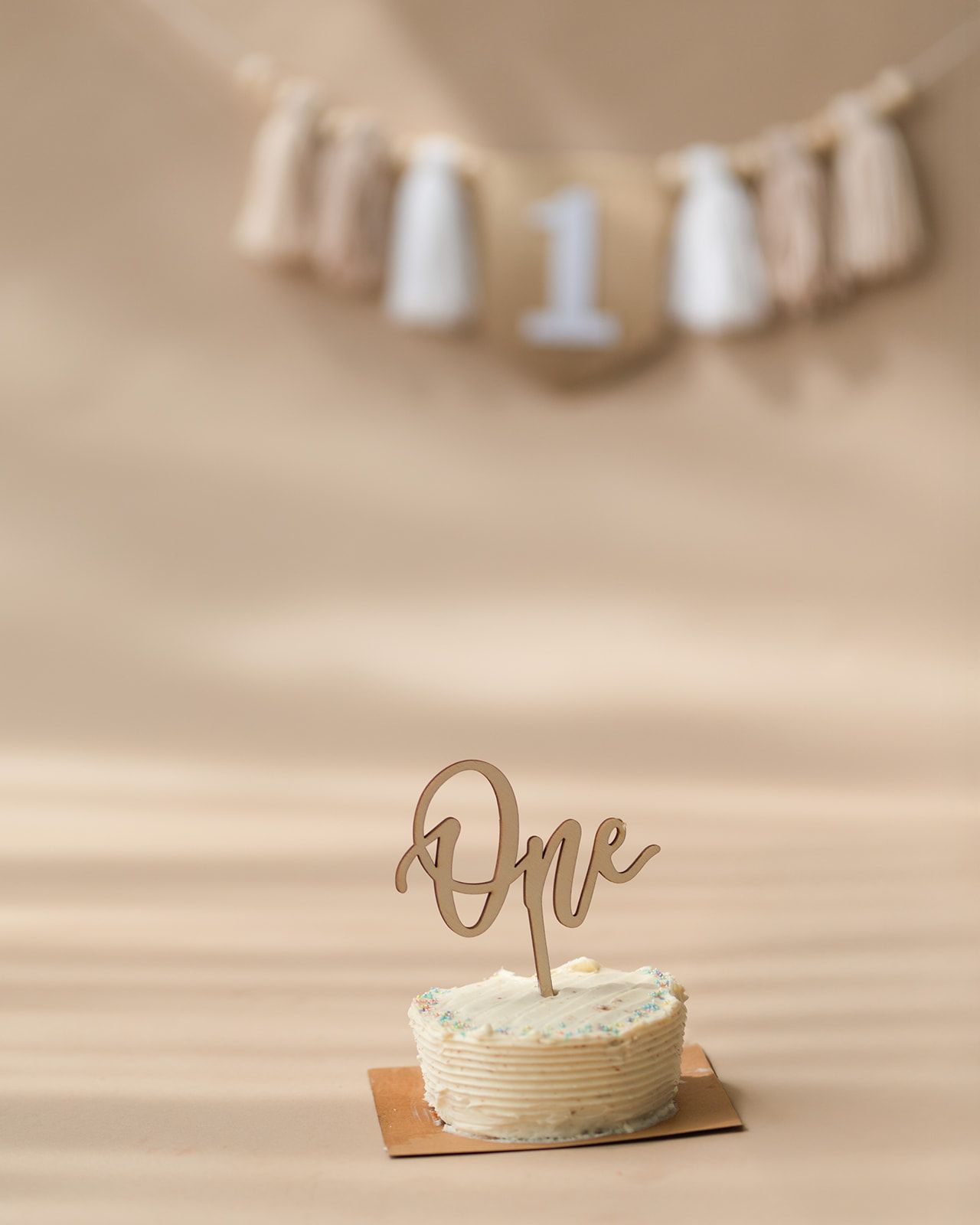 Elegant first birthday smash cake with cursive One topper and boho tassel banner backdrop.