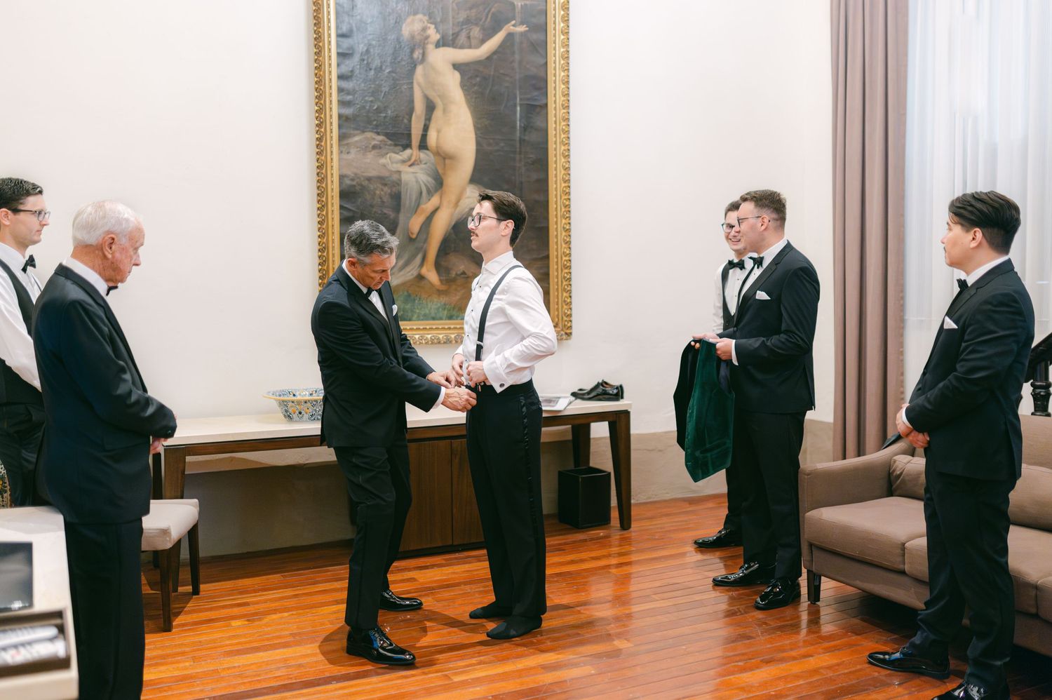 Groom shaking hands with groomsmen in tuxedos before wedding ceremony in elegant room.