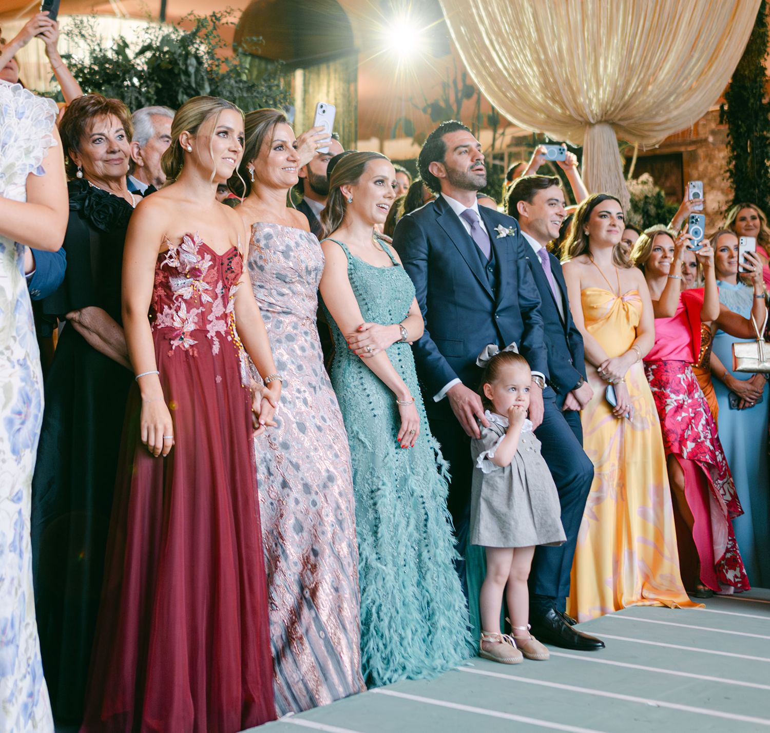 Wedding party lineup in colorful formal gowns with flower girl under decorative floral arch.