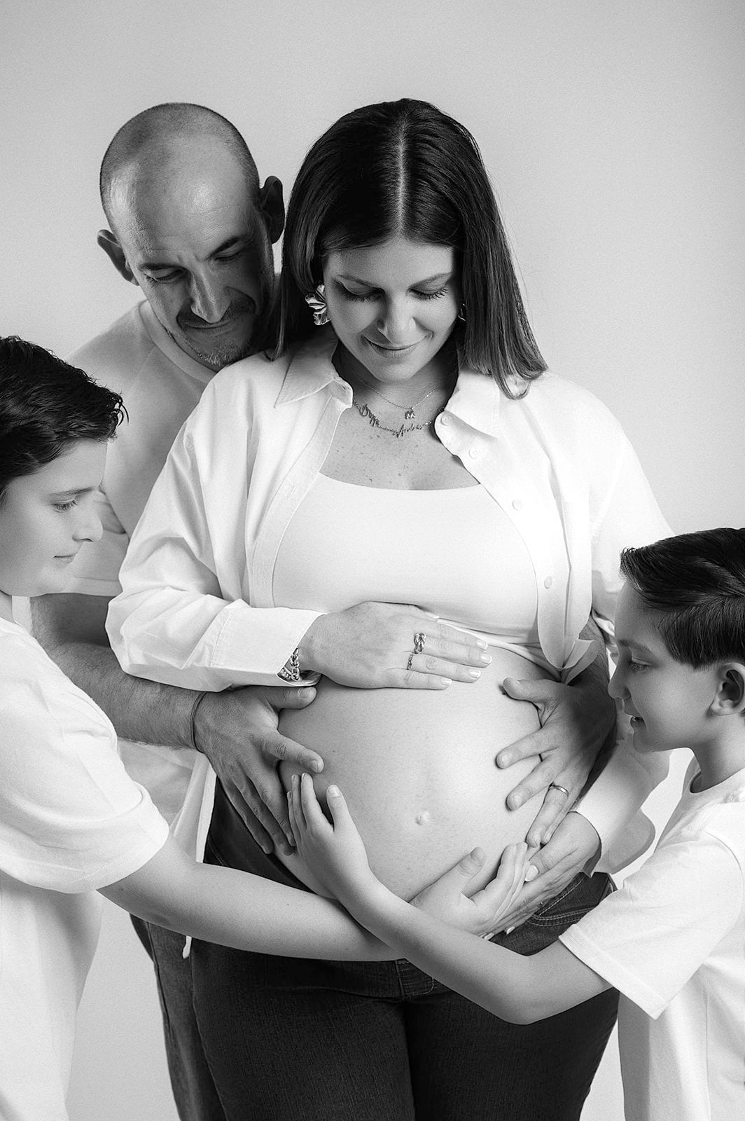 Black and white maternity photo of pregnant woman with husband and two sons tenderly touching her baby bump.