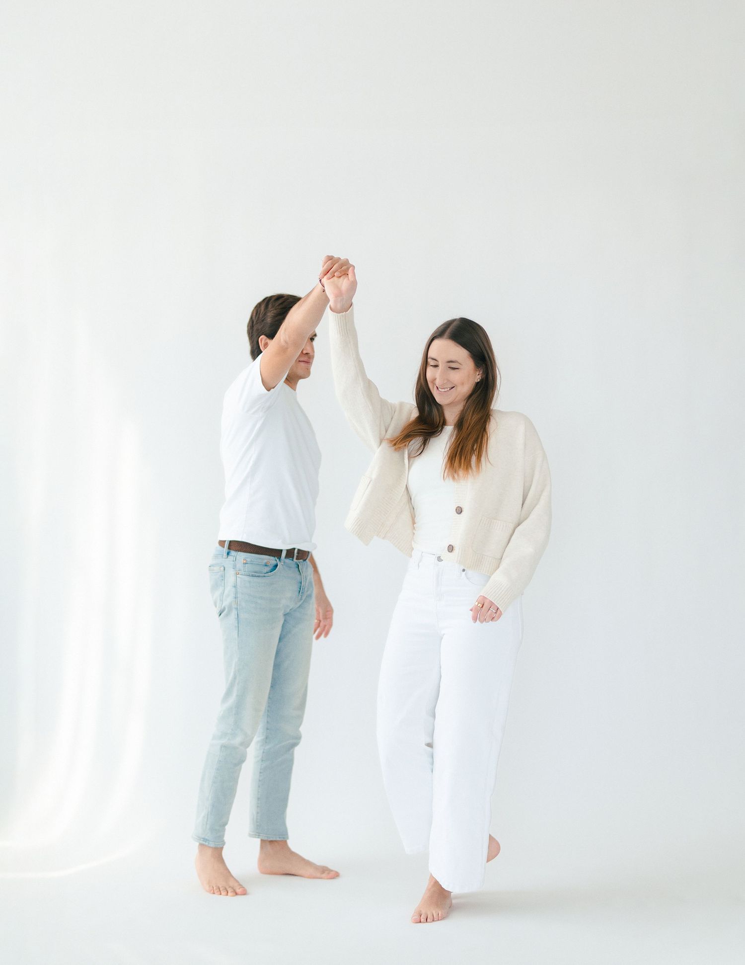 A joyful couple dancing together in a bright white studio, dressed in casual white and cream outfits.