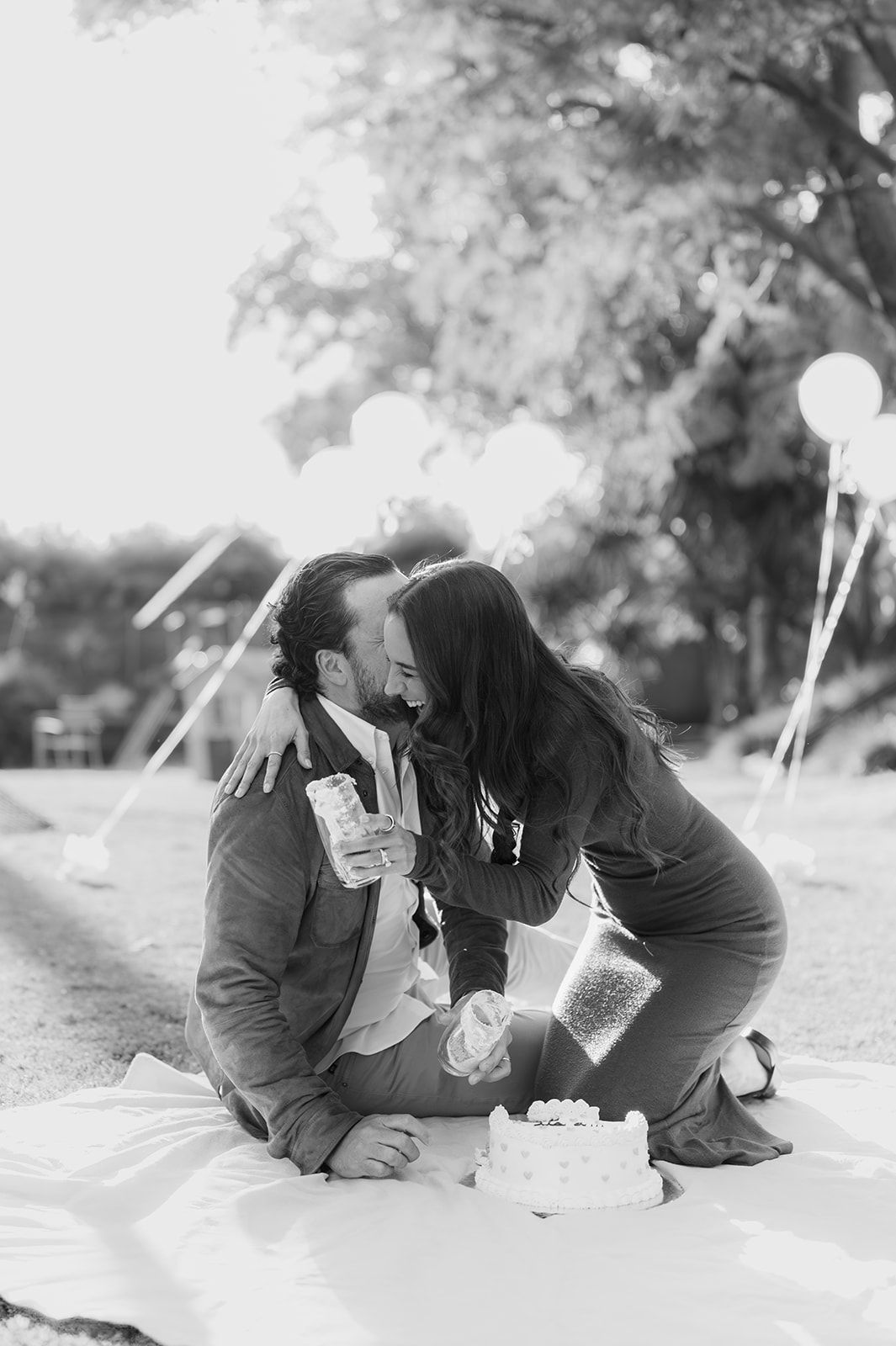 Romantic black and white outdoor photo of a couple sharing a kiss beside a cake at a celebration.