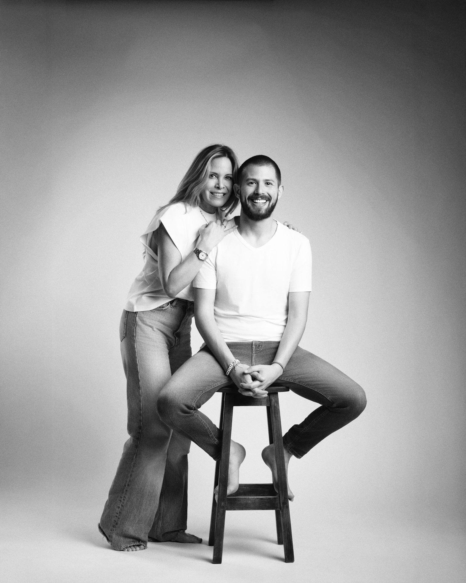Black and white studio portrait of a smiling couple, man seated on stool with woman leaning beside him.