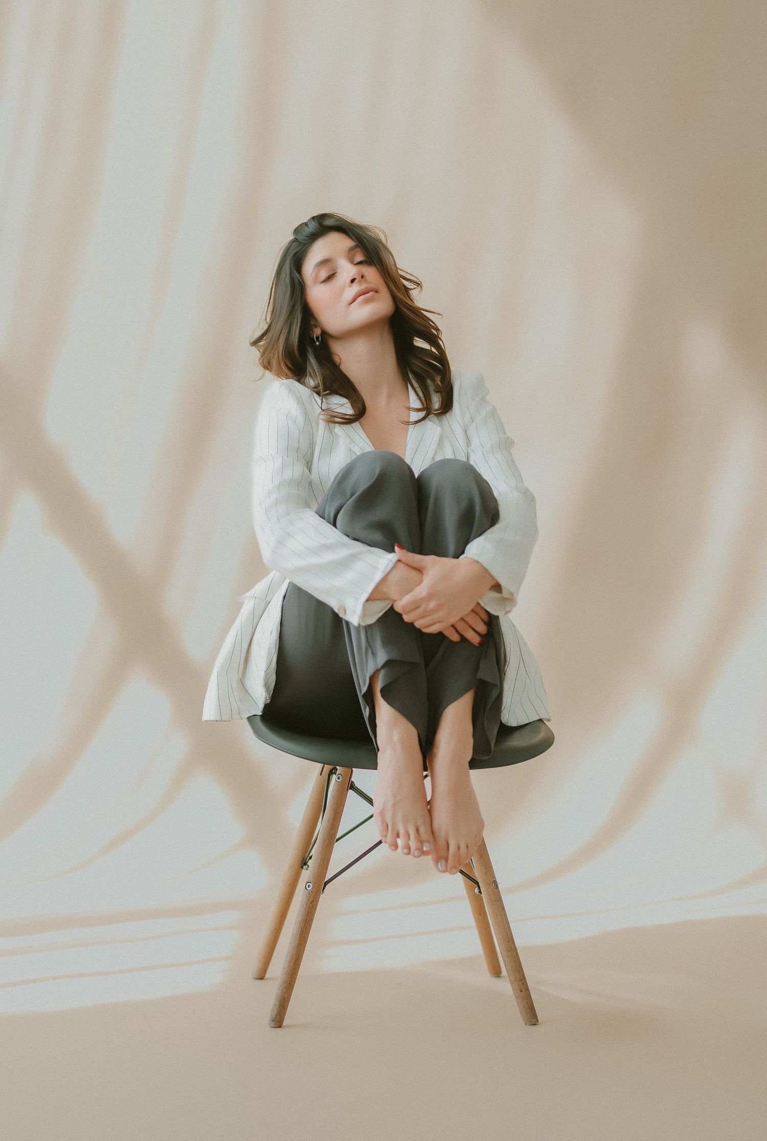 Woman in white blouse and dark pants sits curled on chair with eyes closed, soft draped background.