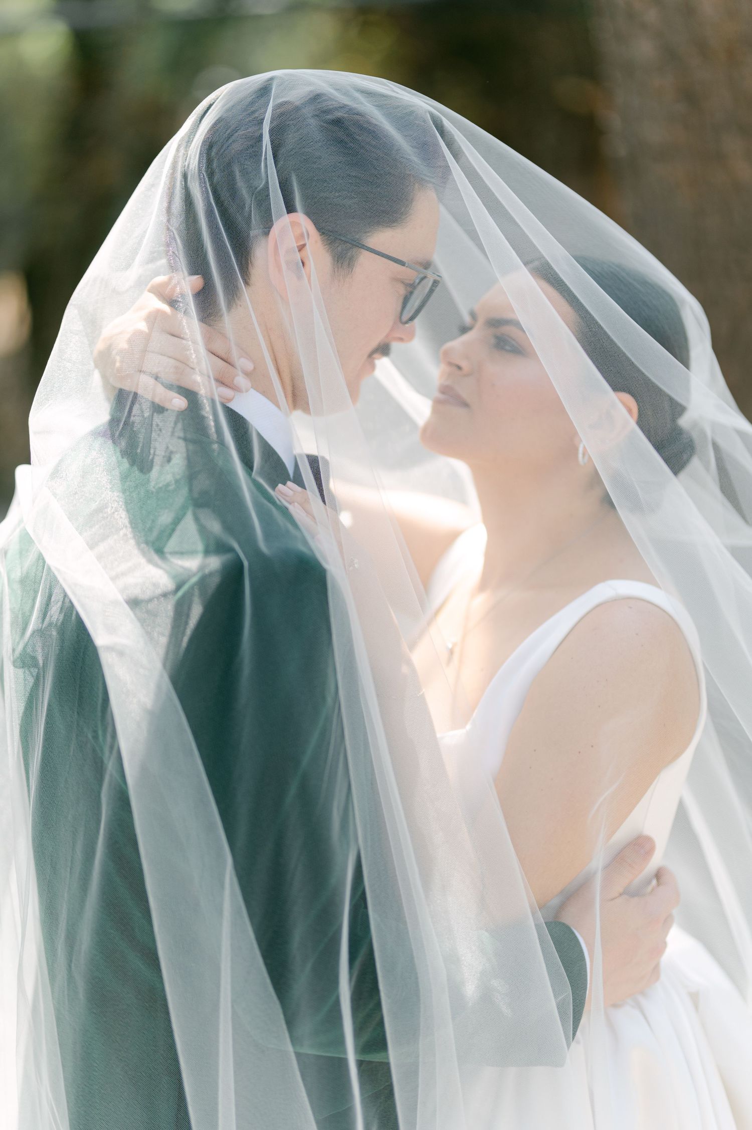 Bride and groom share a romantic moment beneath a flowing veil in a sun-dappled outdoor setting.