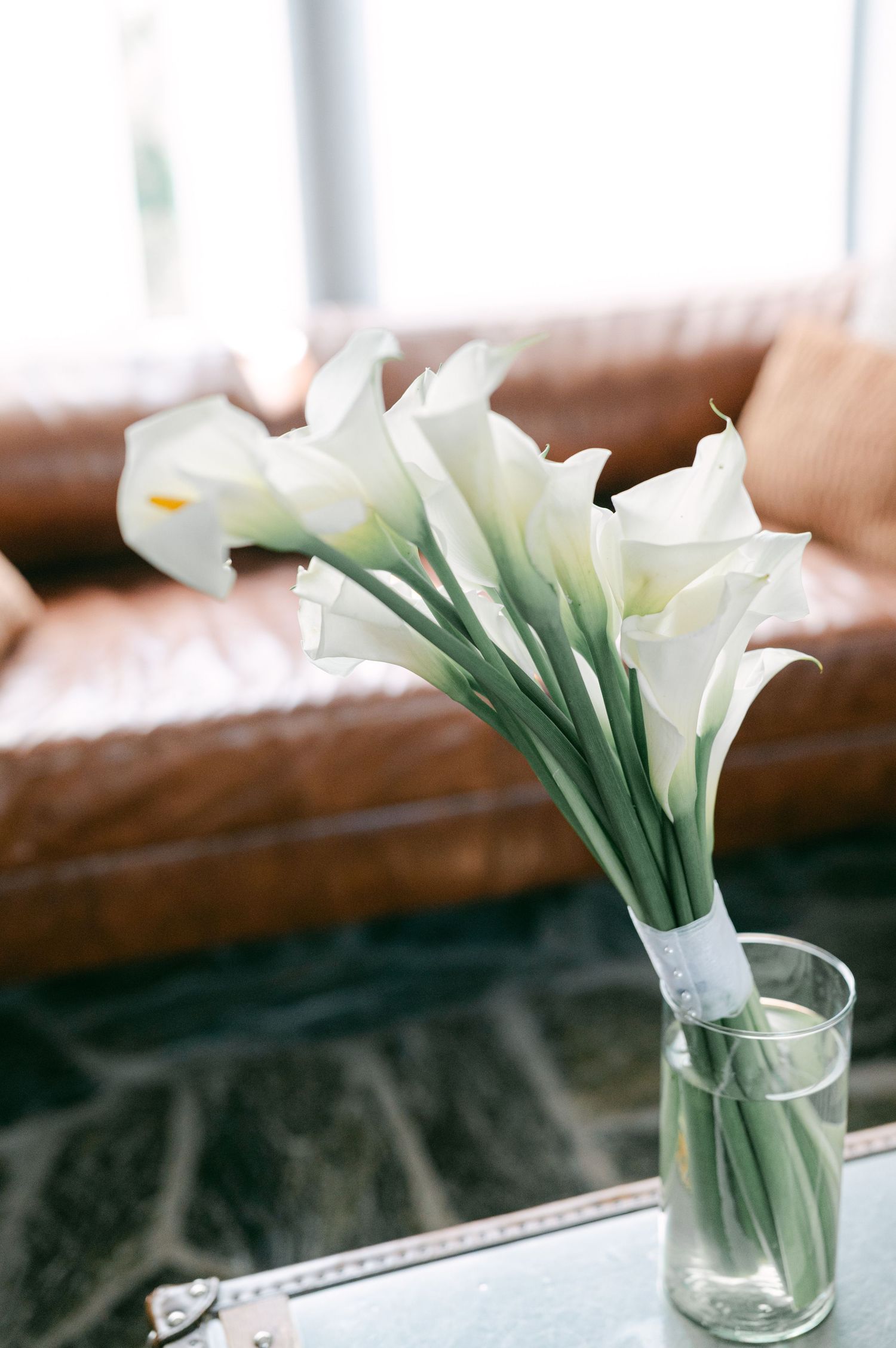 Elegant white calla lily bouquet in a glass vase on a table with leather sofa background.