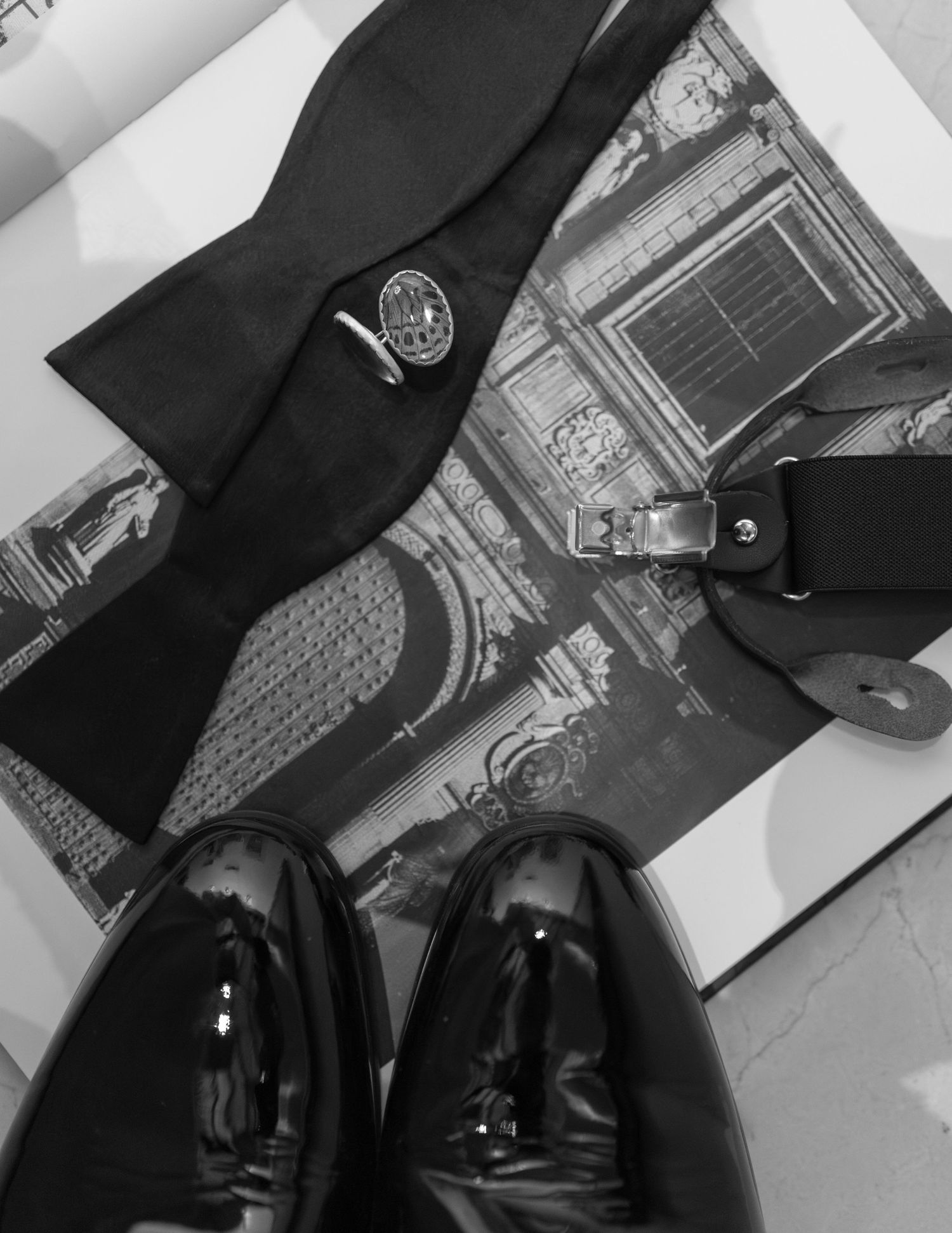 Black and white flat lay of groom wedding accessories including bow tie, cufflinks, belt, and patent shoes.