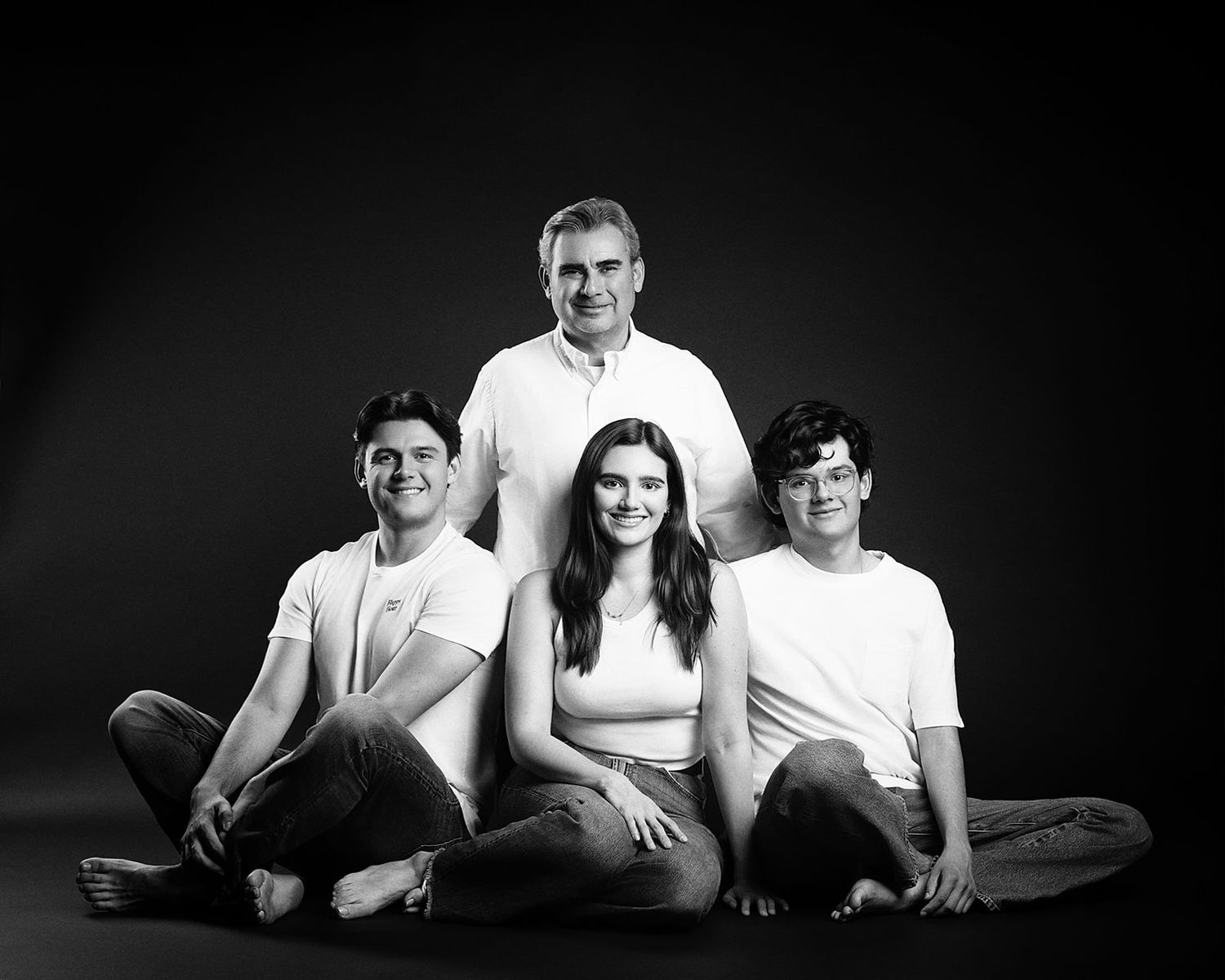 Black and white family studio portrait of a father standing behind three smiling teenagers on the floor.