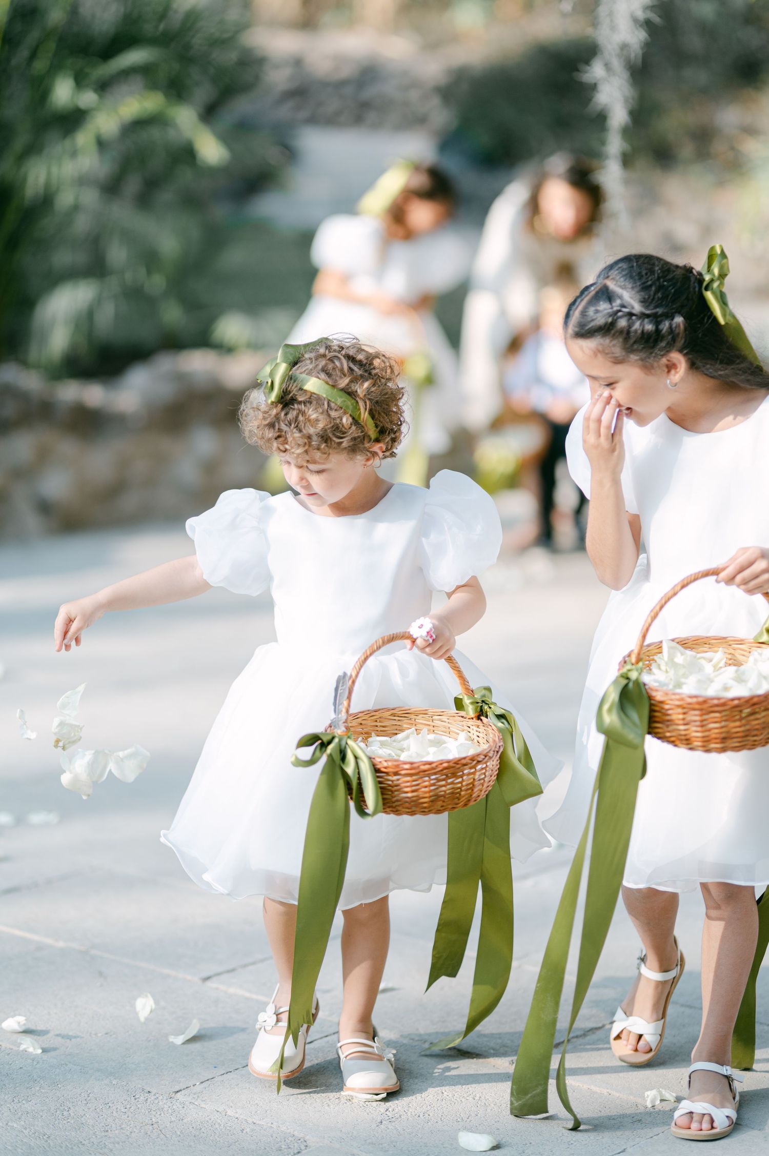 Flower girls in white dresses with green ribbon baskets scattering petals at an outdoor wedding ceremony.
