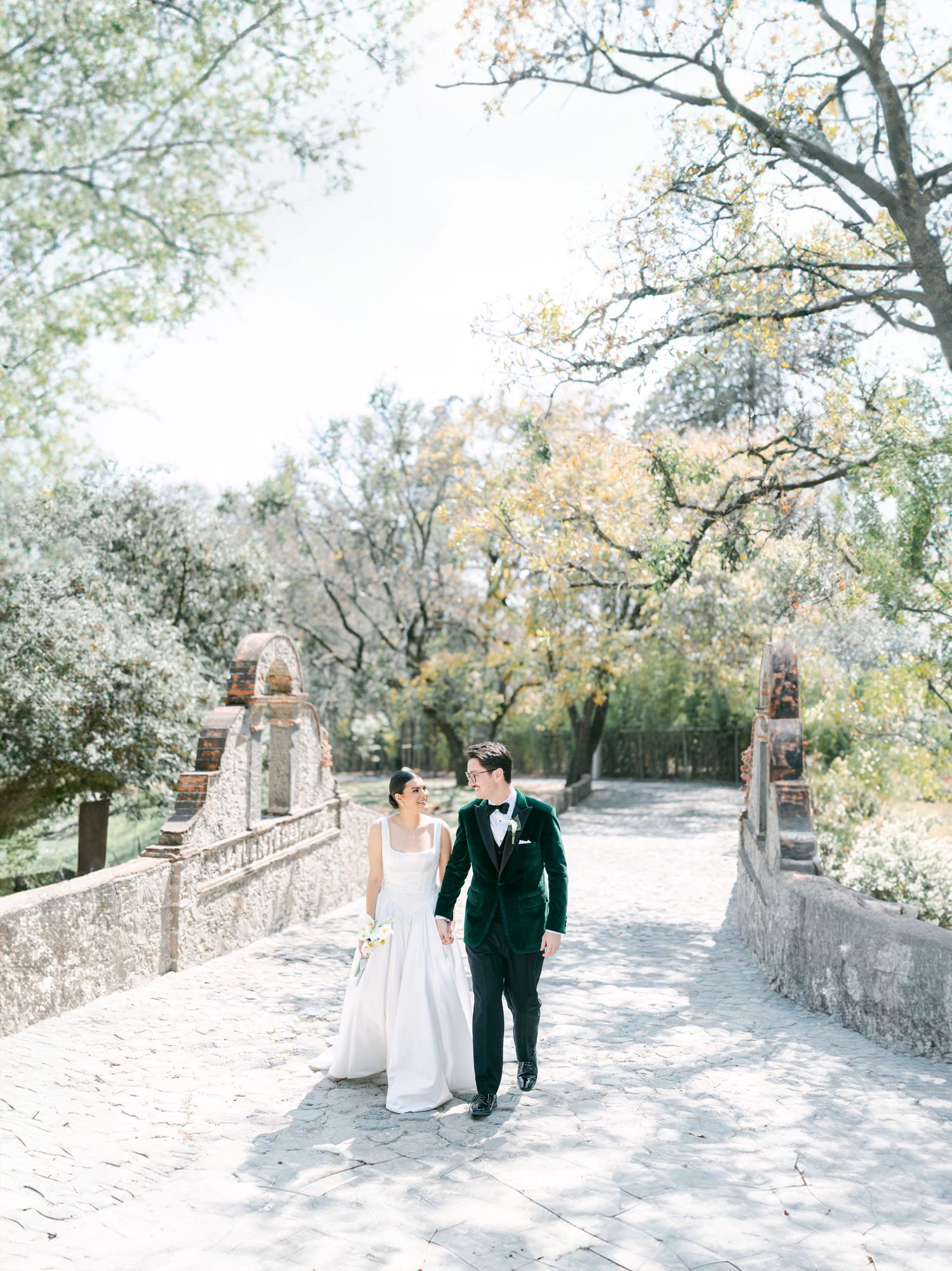 Bride and groom walking hand in hand along a stone path surrounded by autumn trees.