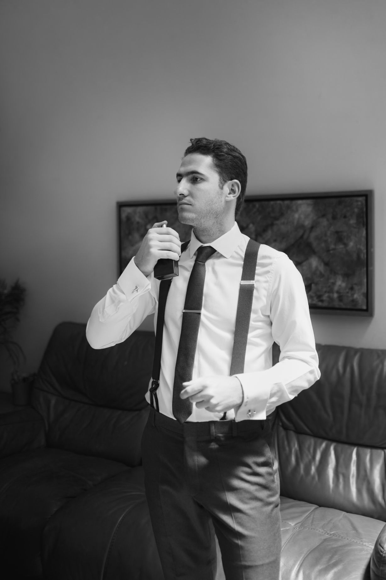 Black and white photo of groomsman in white shirt and suspenders holding tie before wedding.