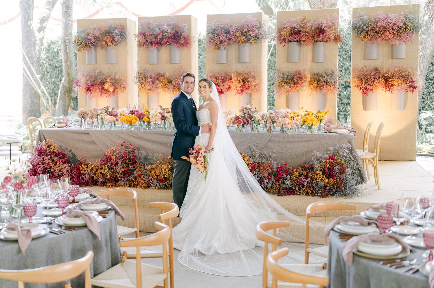 Bride and groom stand before a stunning colorful floral backdrop wall at elegant wedding reception.