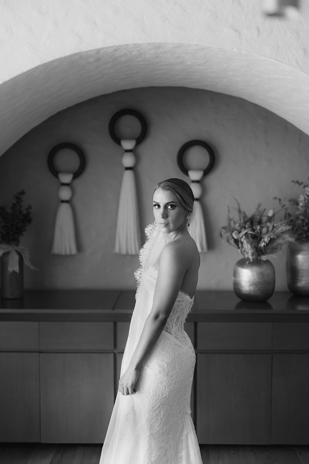 Bride in lace wedding gown poses under arched doorway in black and white portrait.