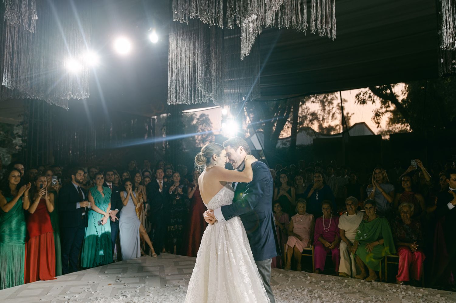 Bride in white gown dances under spotlights with guests watching in colorful attire.