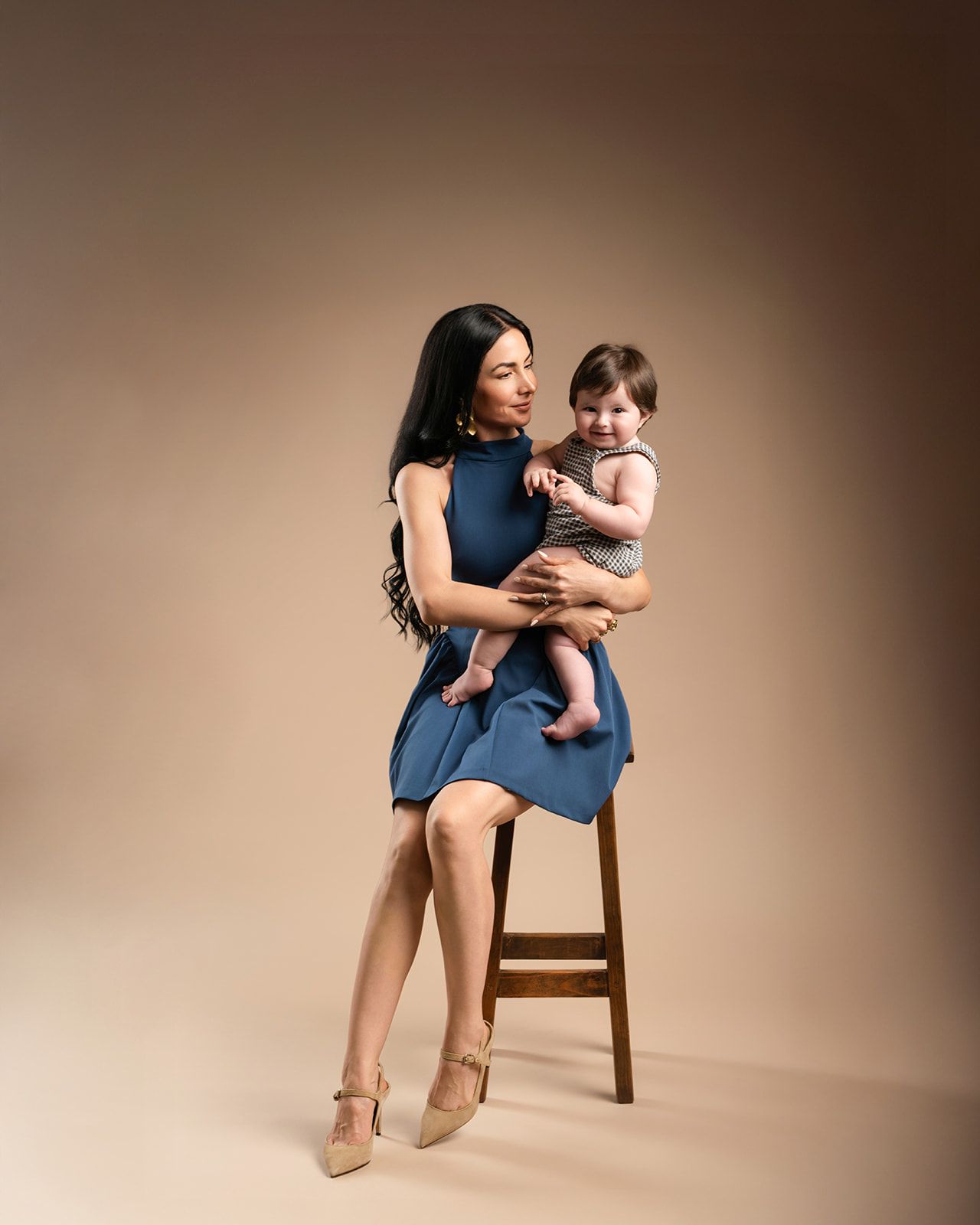 A stylish mother in a blue dress holds her baby while seated on a stool in a warm-toned studio portrait.