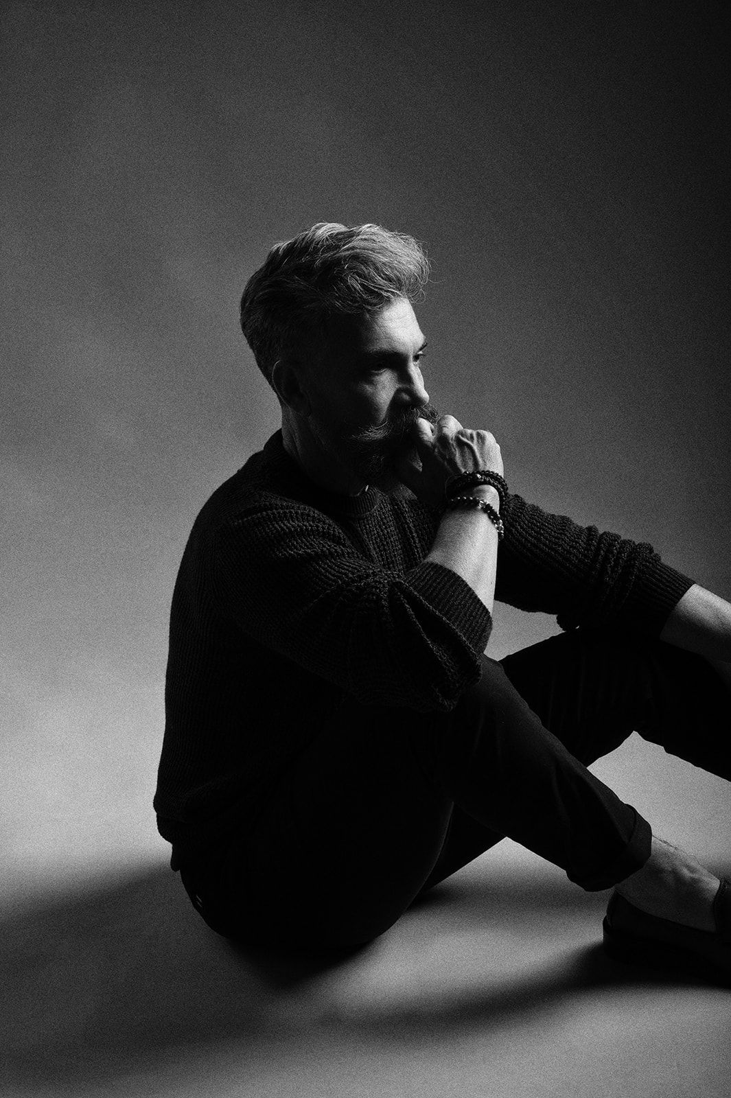 Moody black and white portrait of a young man sitting on floor in dark sweater, contemplating.