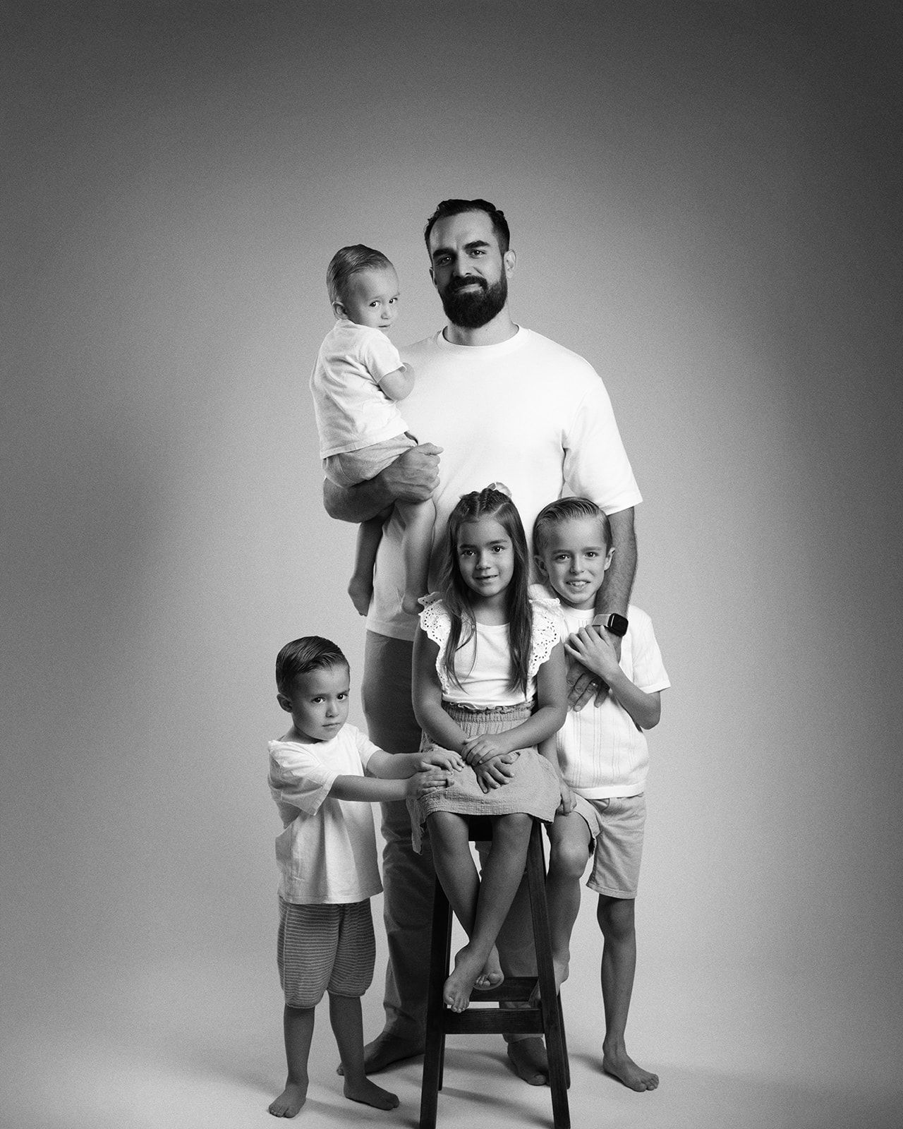 Black and white studio portrait of a bearded father standing with four children around a stool.