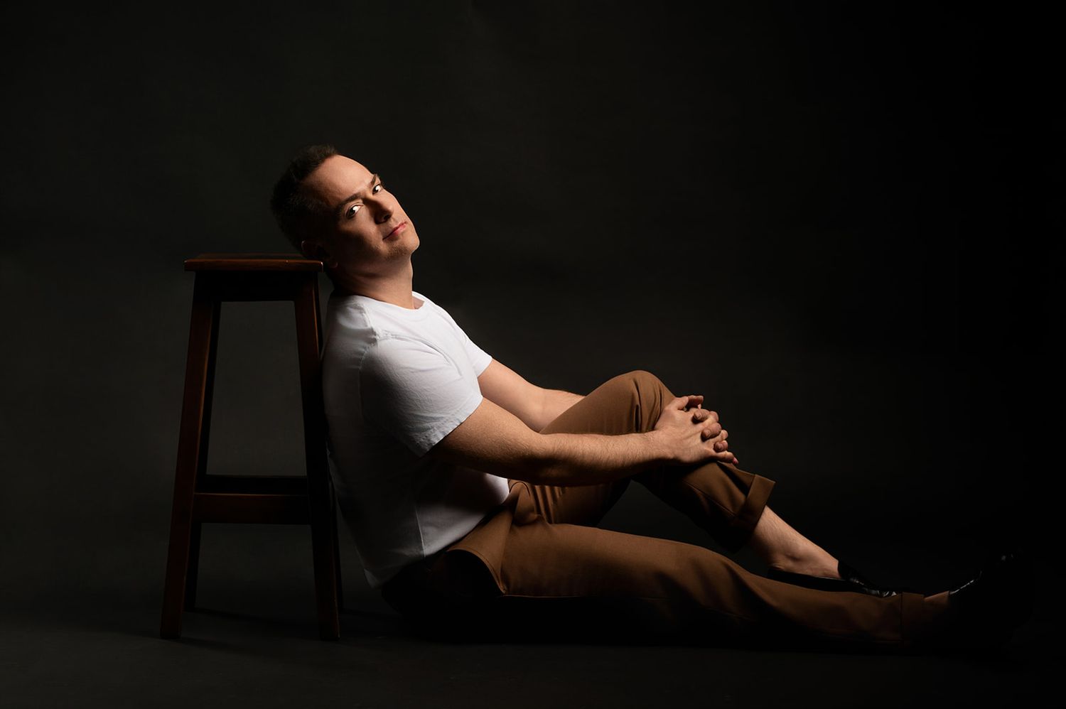 Man in white t-shirt and brown pants relaxing on floor beside wooden stool, studio portrait.