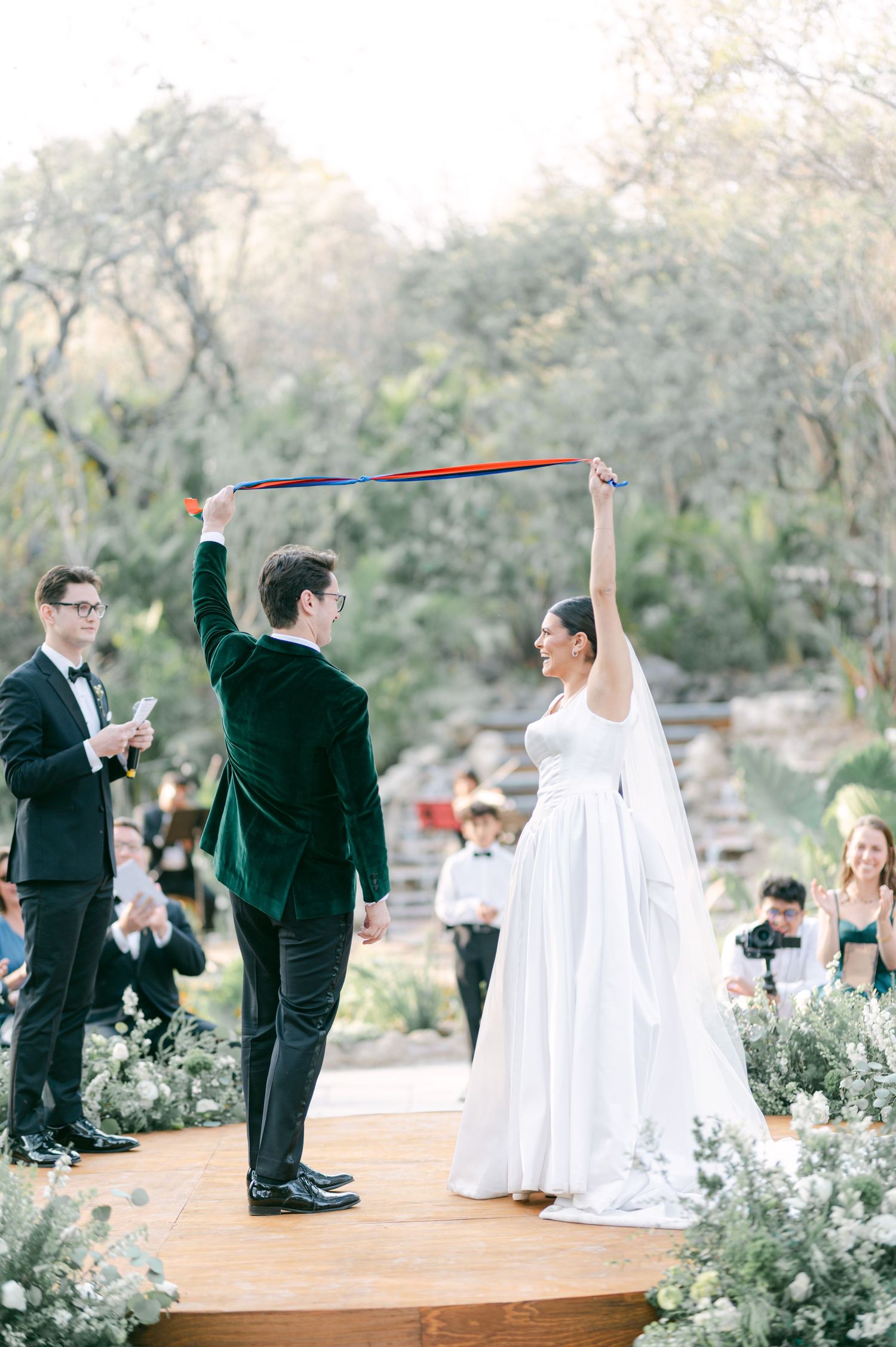 Bride and groom celebrate at outdoor ceremony holding colorful ribbon while guests cheer in wooded setting.