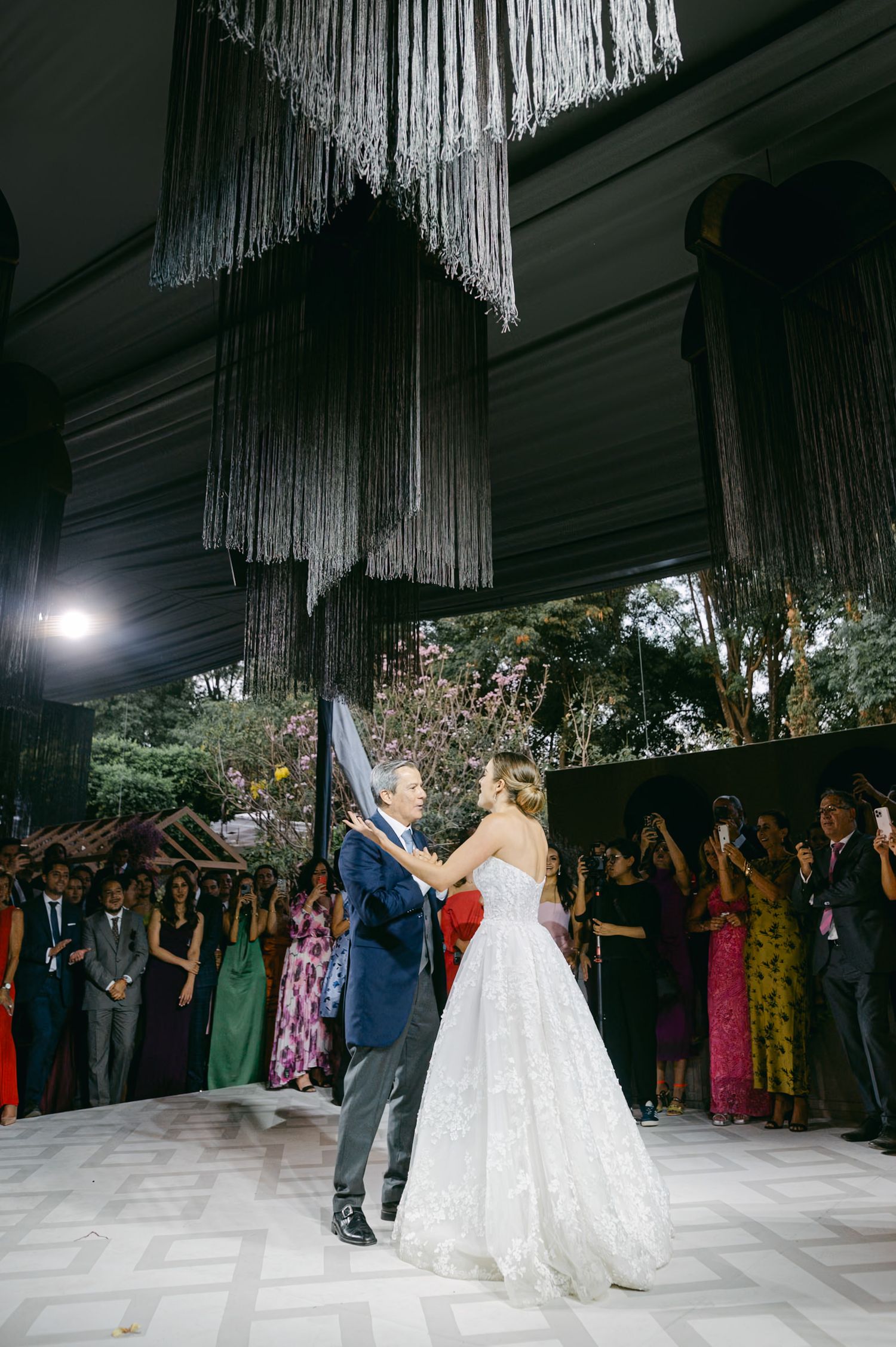 Newlyweds share their first dance under dramatic fringe chandeliers while guests watch from the sides.