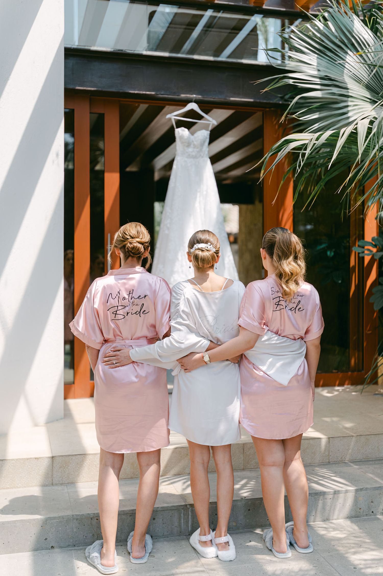 A bride and two women in pink robes labeled Mother of Bride stand before a hanging wedding gown.