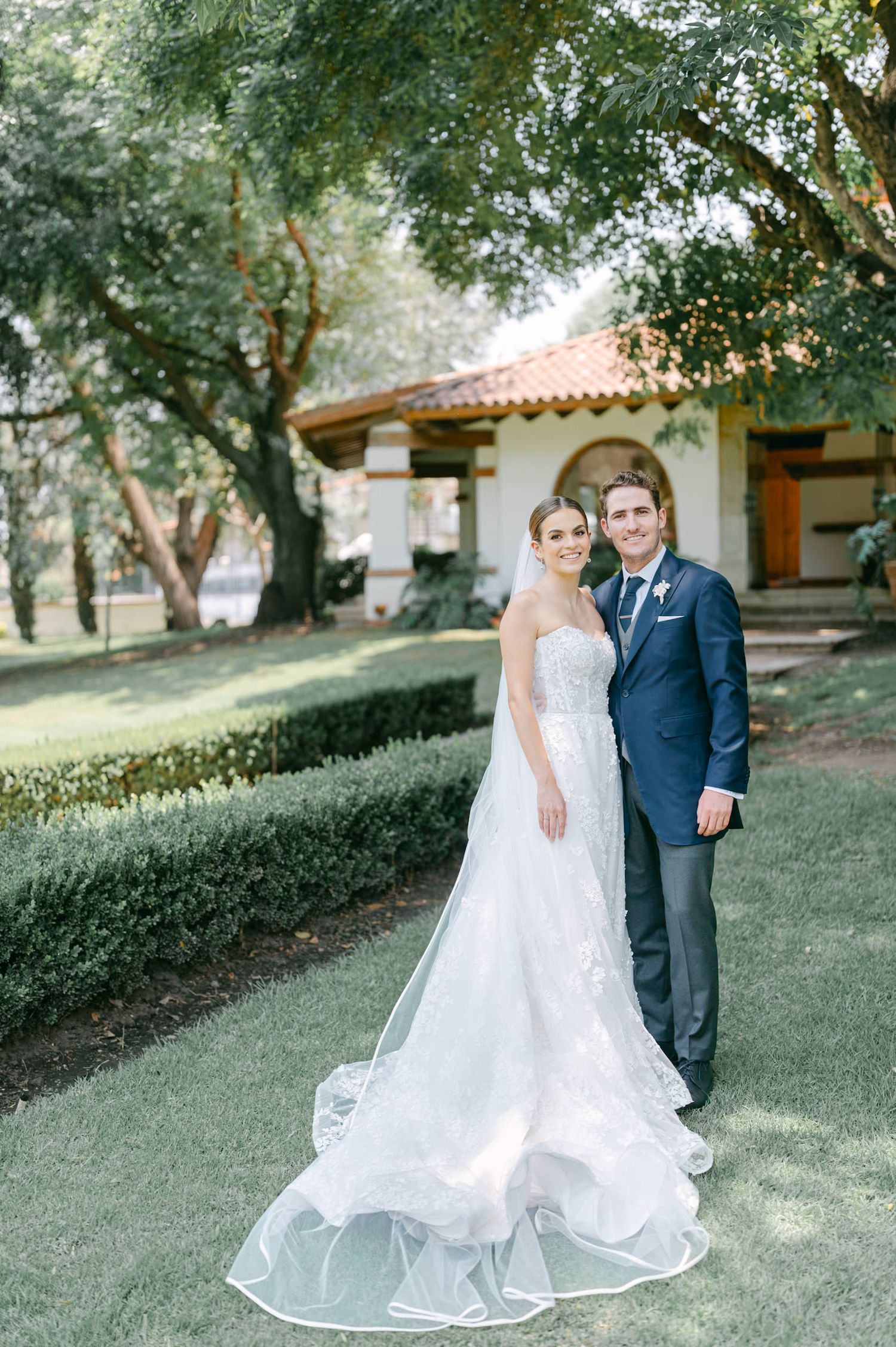 Bride in lace gown and groom in navy suit pose together on green lawn before a Spanish-style venue.