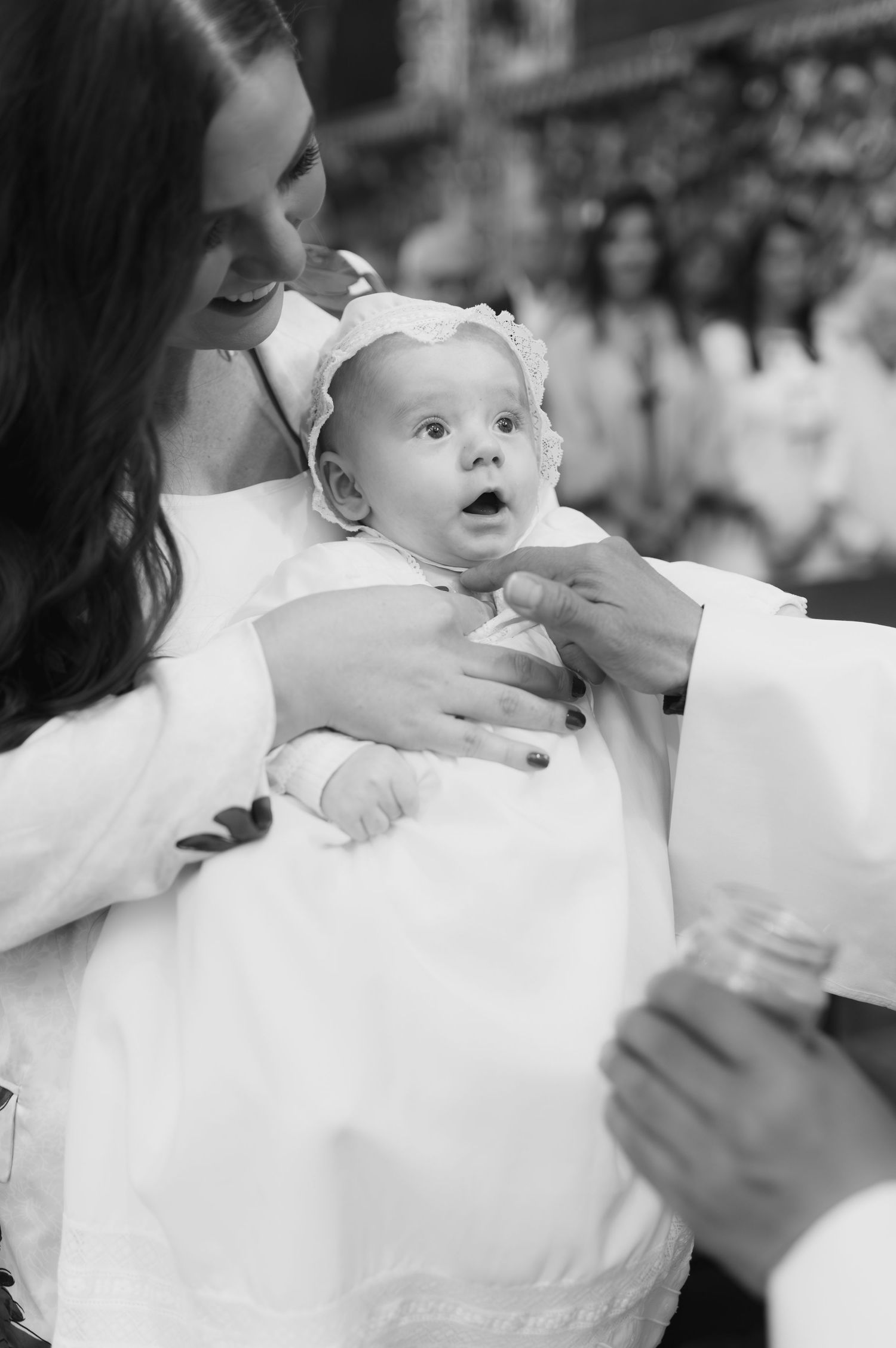 Black and white photo of a baby in a white bonnet being held at a baptism ceremony.