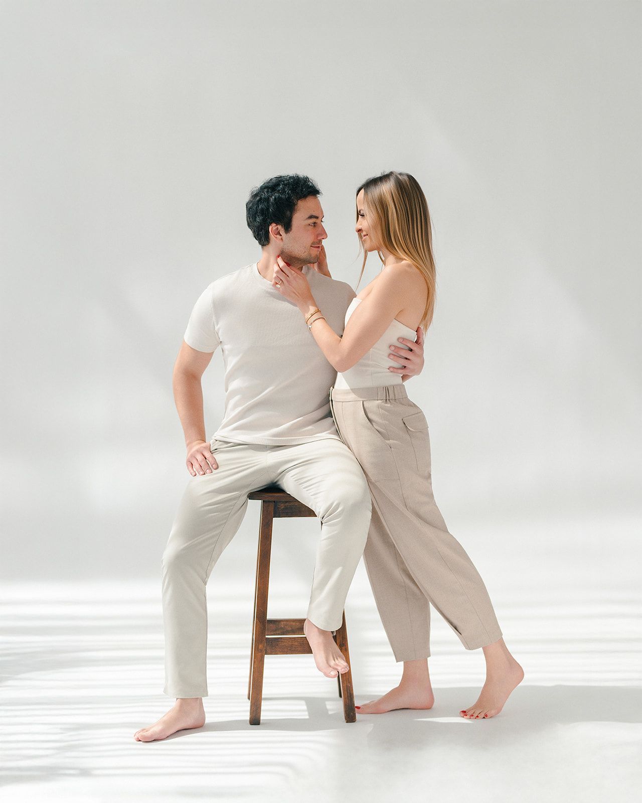 Couple in matching neutral beige outfits posing intimately on a wooden stool against a white background.