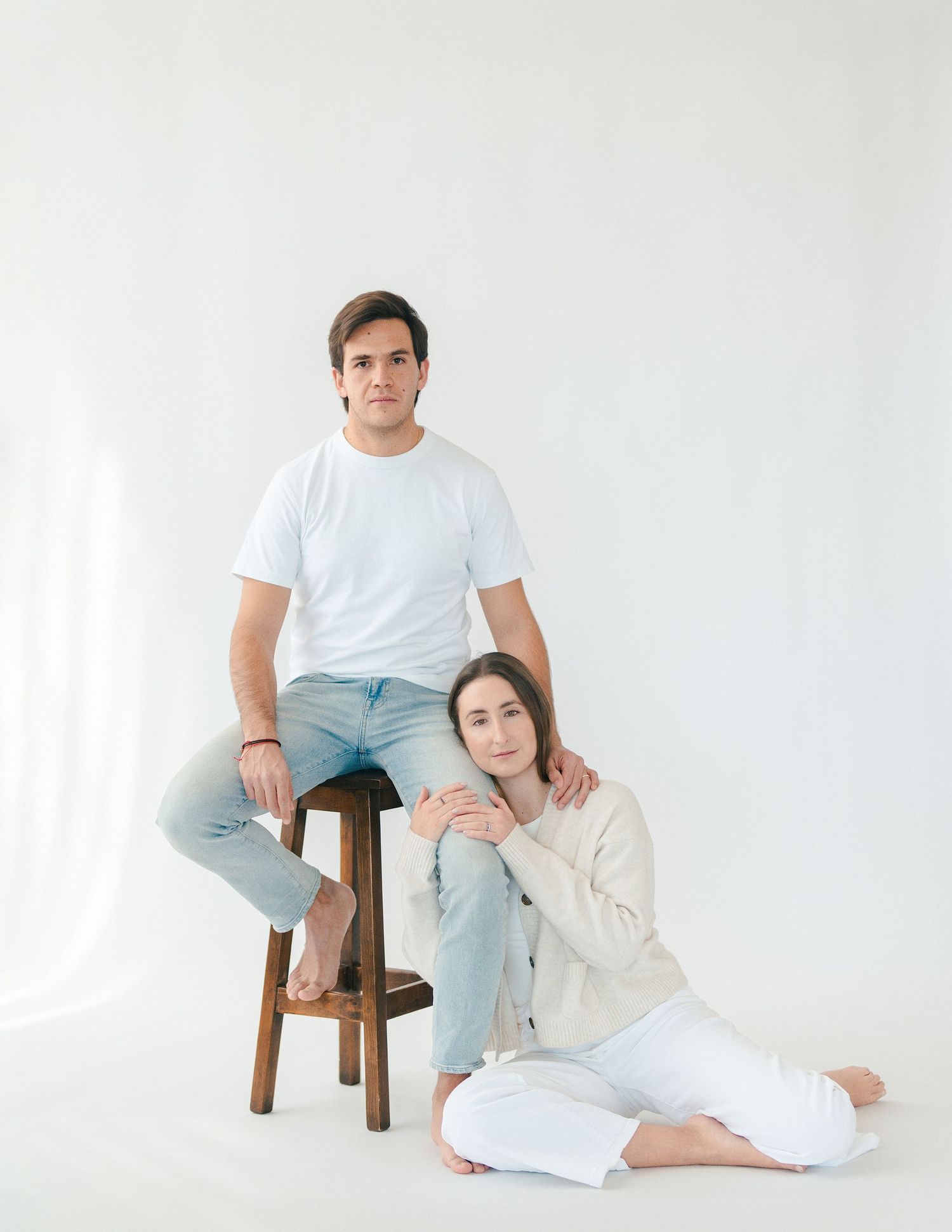 A couple posing elegantly in a white studio, man seated on a stool and woman sitting on the floor.