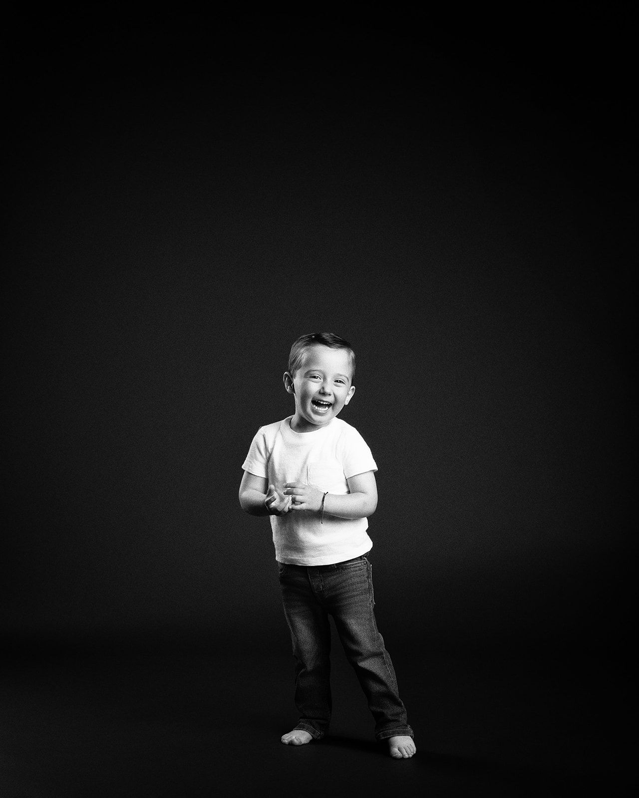 Laughing young boy in white t-shirt and jeans poses barefoot against dark studio background.
