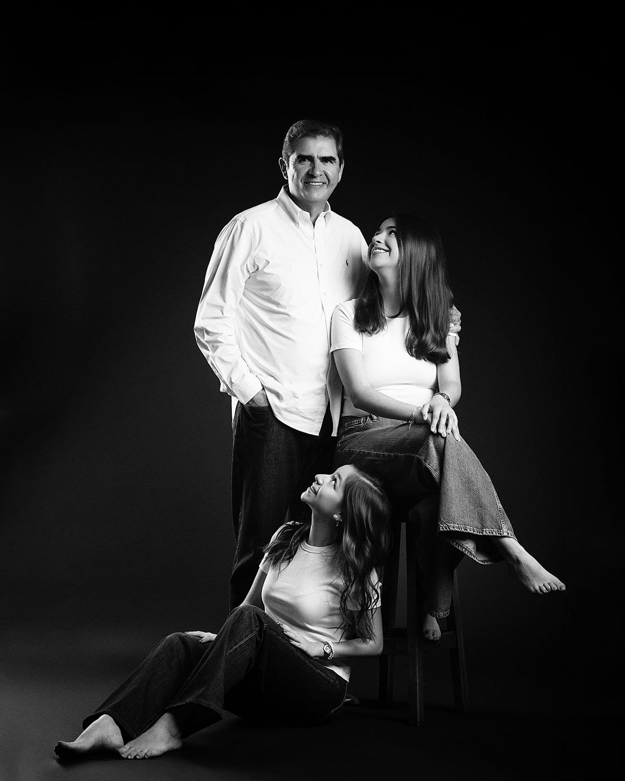 Black and white studio portrait of a man standing with two young women posed around a stool.