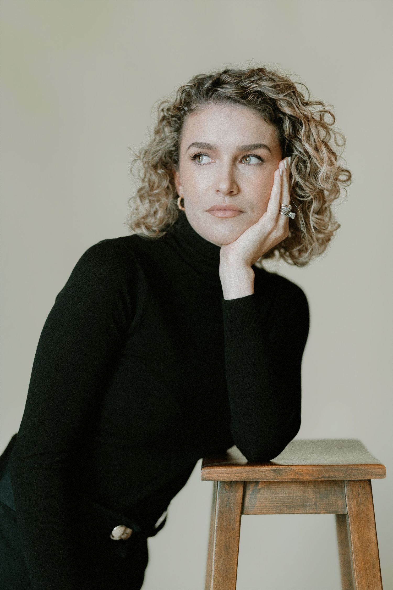 Curly-haired woman in black turtleneck leaning on wooden stool, looking up, studio portrait.