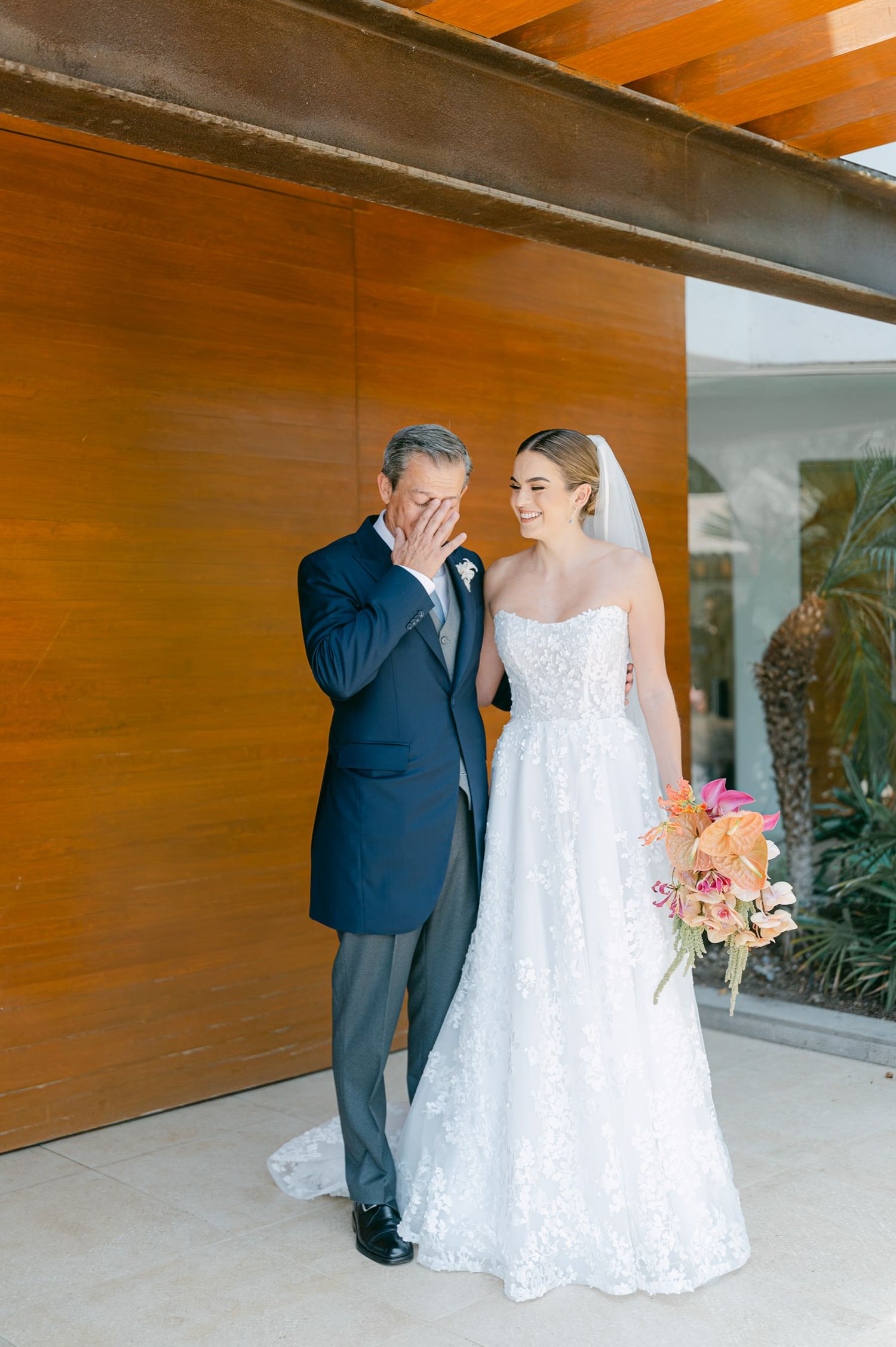 Emotional father wiping tears beside smiling bride in lace wedding dress holding colorful bridal bouquet.