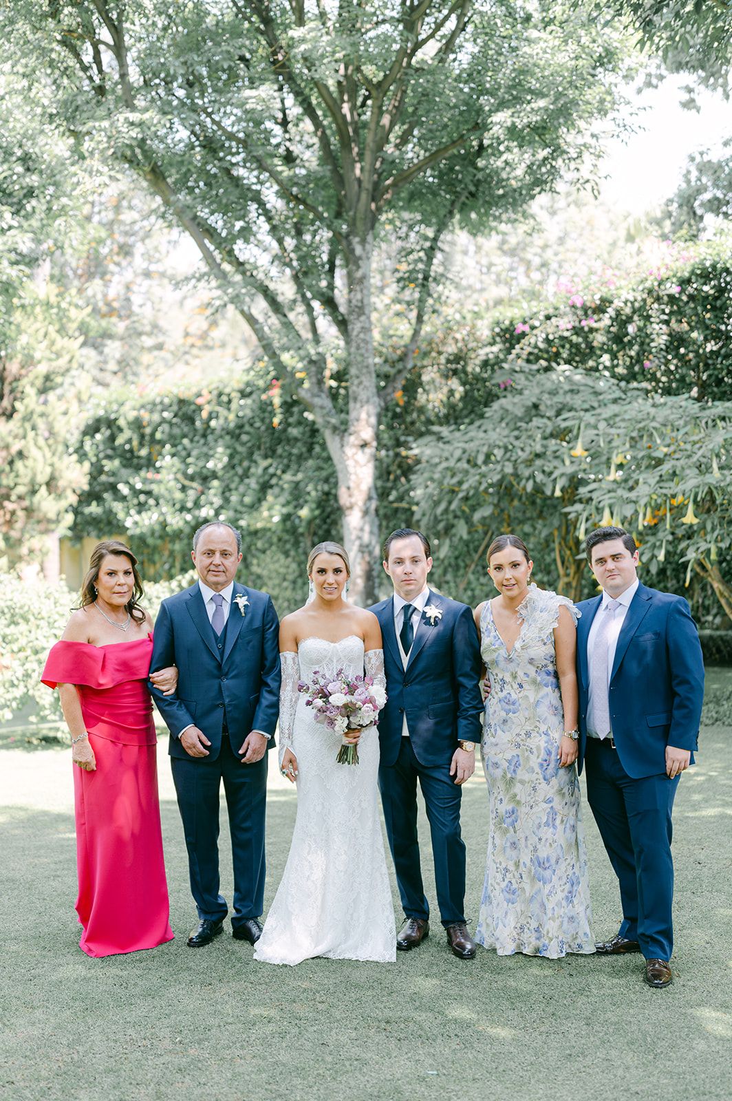 Bride and groom pose with family of six in garden, wearing elegant formal attire in blue and floral gowns.