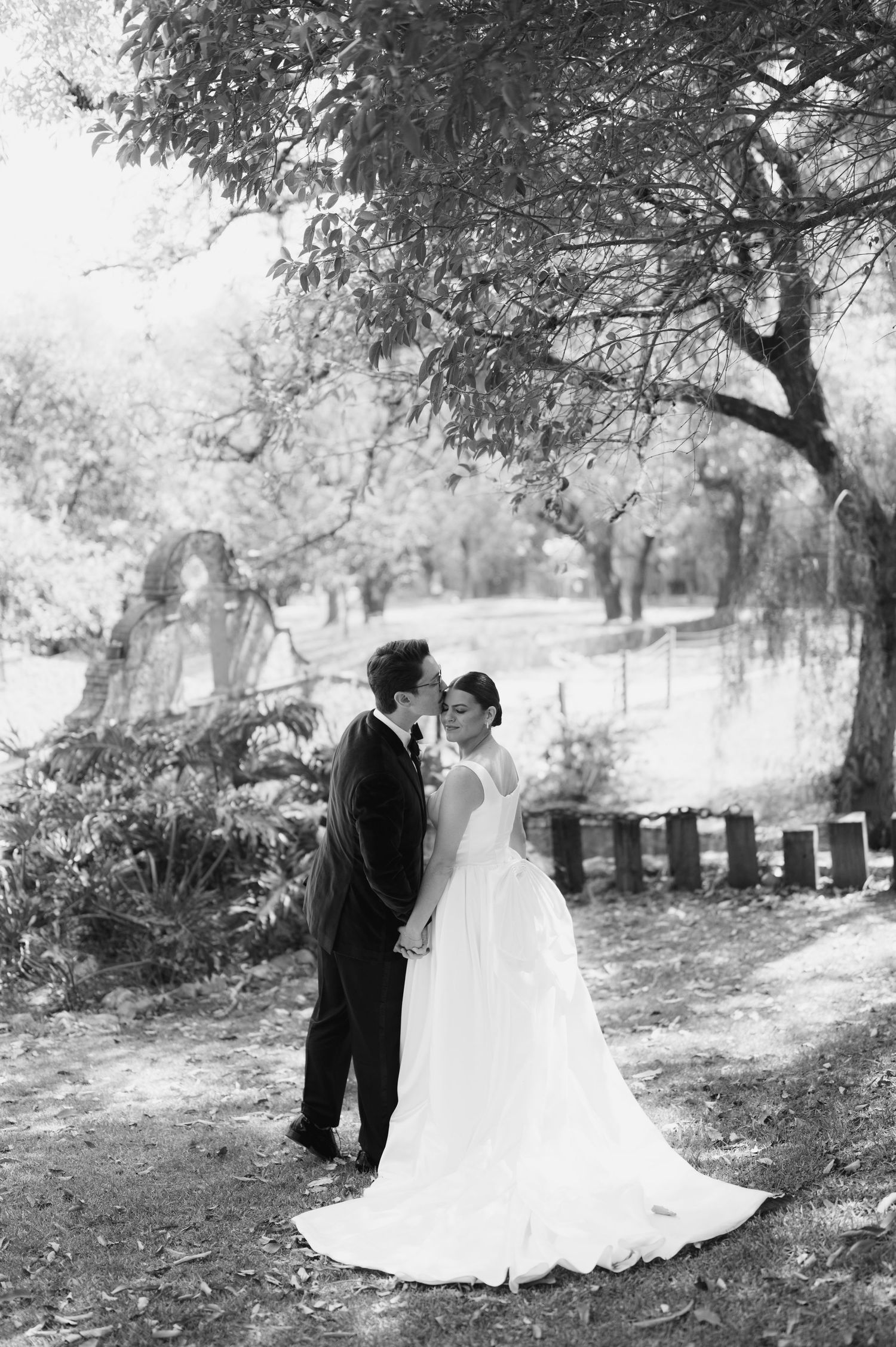 Black and white romantic portrait of bride and groom embracing tenderly under a large tree in garden setting.