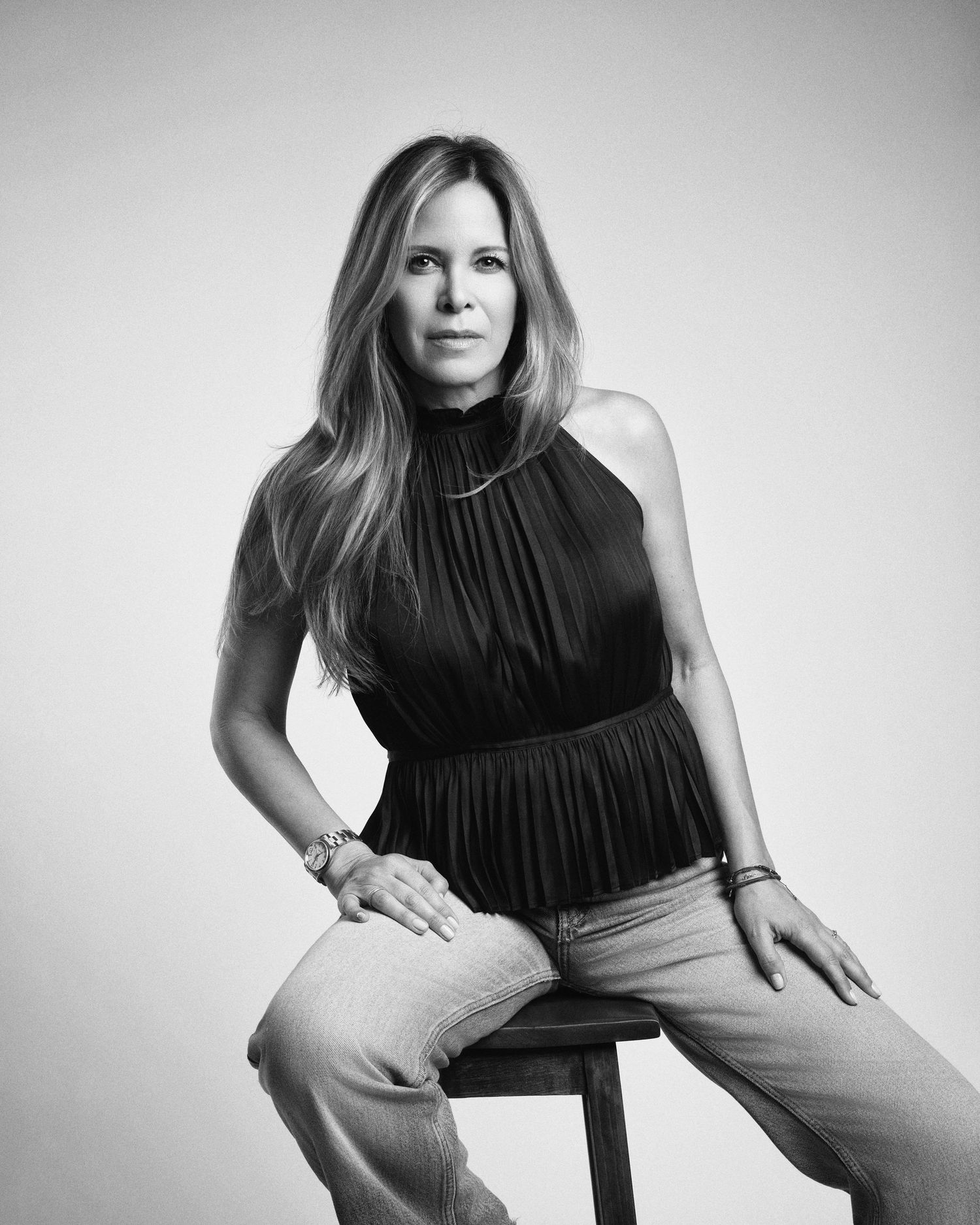 Black and white solo portrait of a woman in a black halter top and jeans seated on a wooden stool.