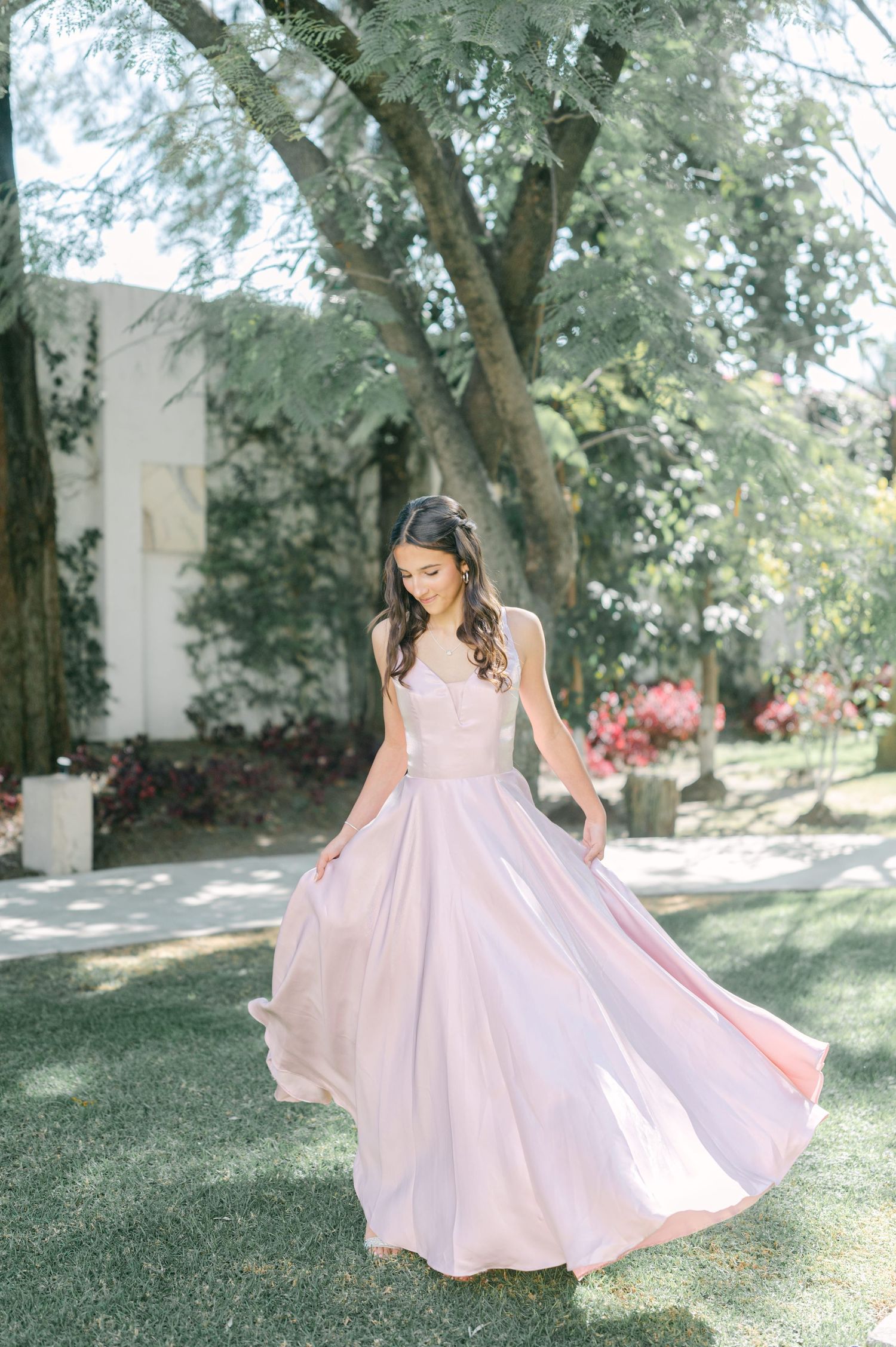 A bride in a flowing pink gown twirls in a sun-drenched garden setting.
