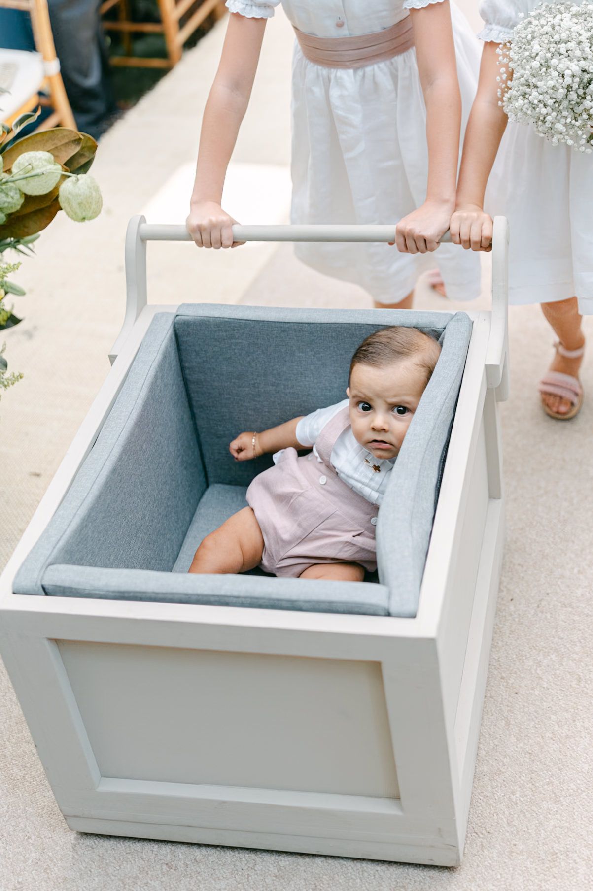 Adorable baby in pink outfit sits in a padded gray cart being pushed down the wedding ceremony aisle.