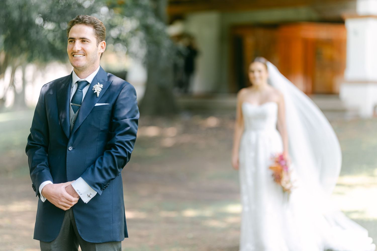Groom in navy suit waiting for first look as bride in lace gown approaches from behind.