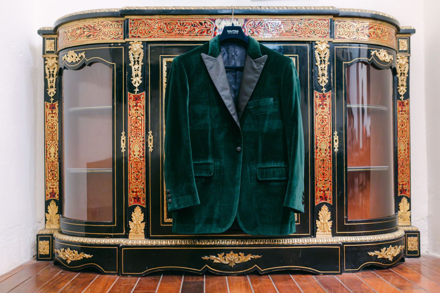 Emerald green velvet wedding blazer hanging on ornate antique cabinet with gold details.