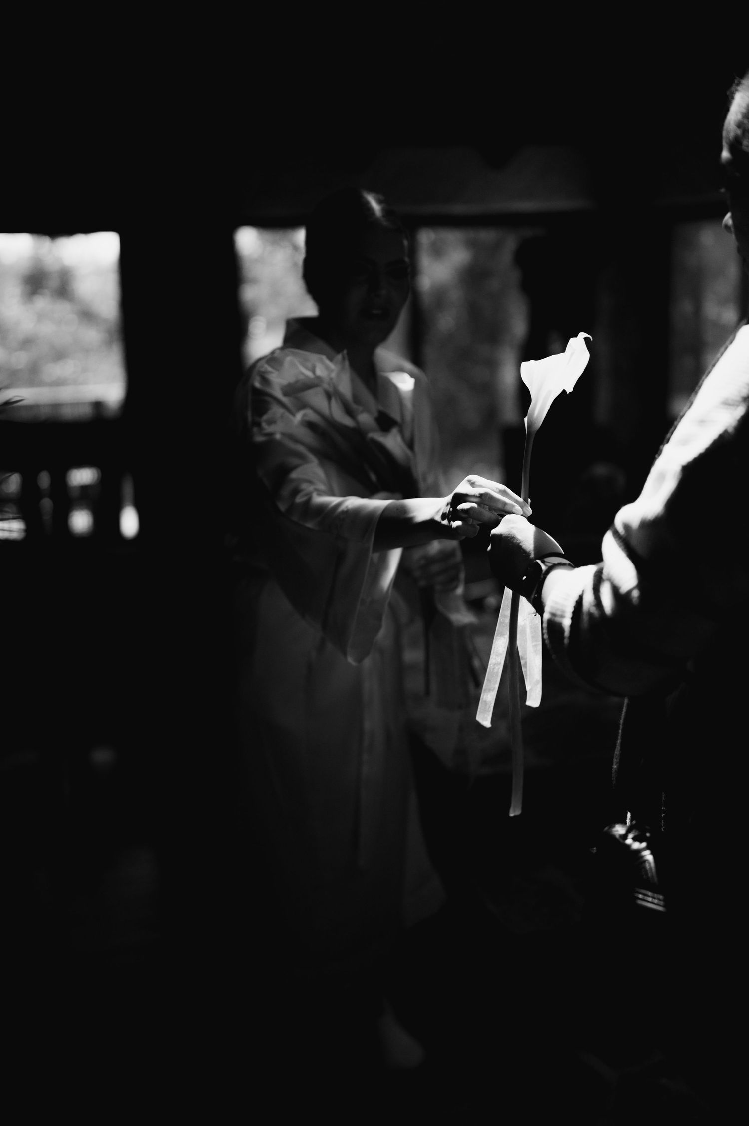 Dramatic black and white image of bride and groom holding a single calla lily in a dimly lit indoor setting.
