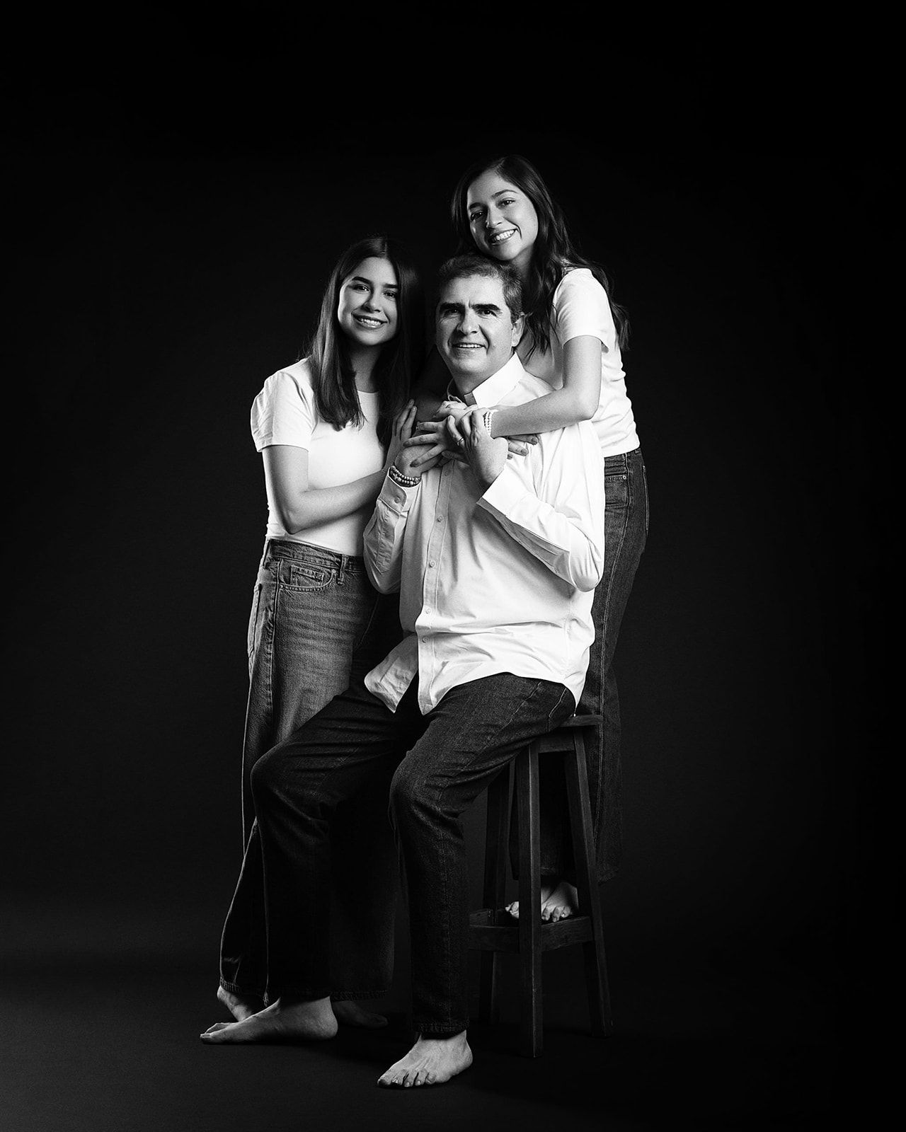 Black and white studio portrait of a man seated on a stool with two smiling young women beside him.